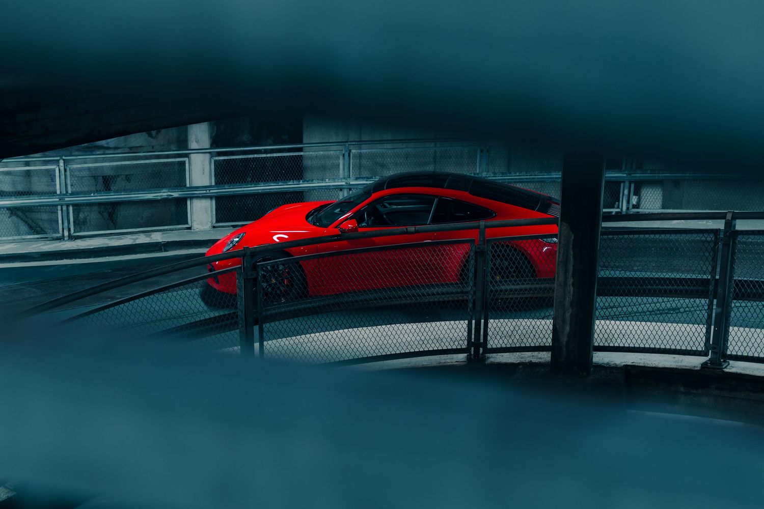 Red sports car photographed through parking garage architecture creating a moody atmosphere.