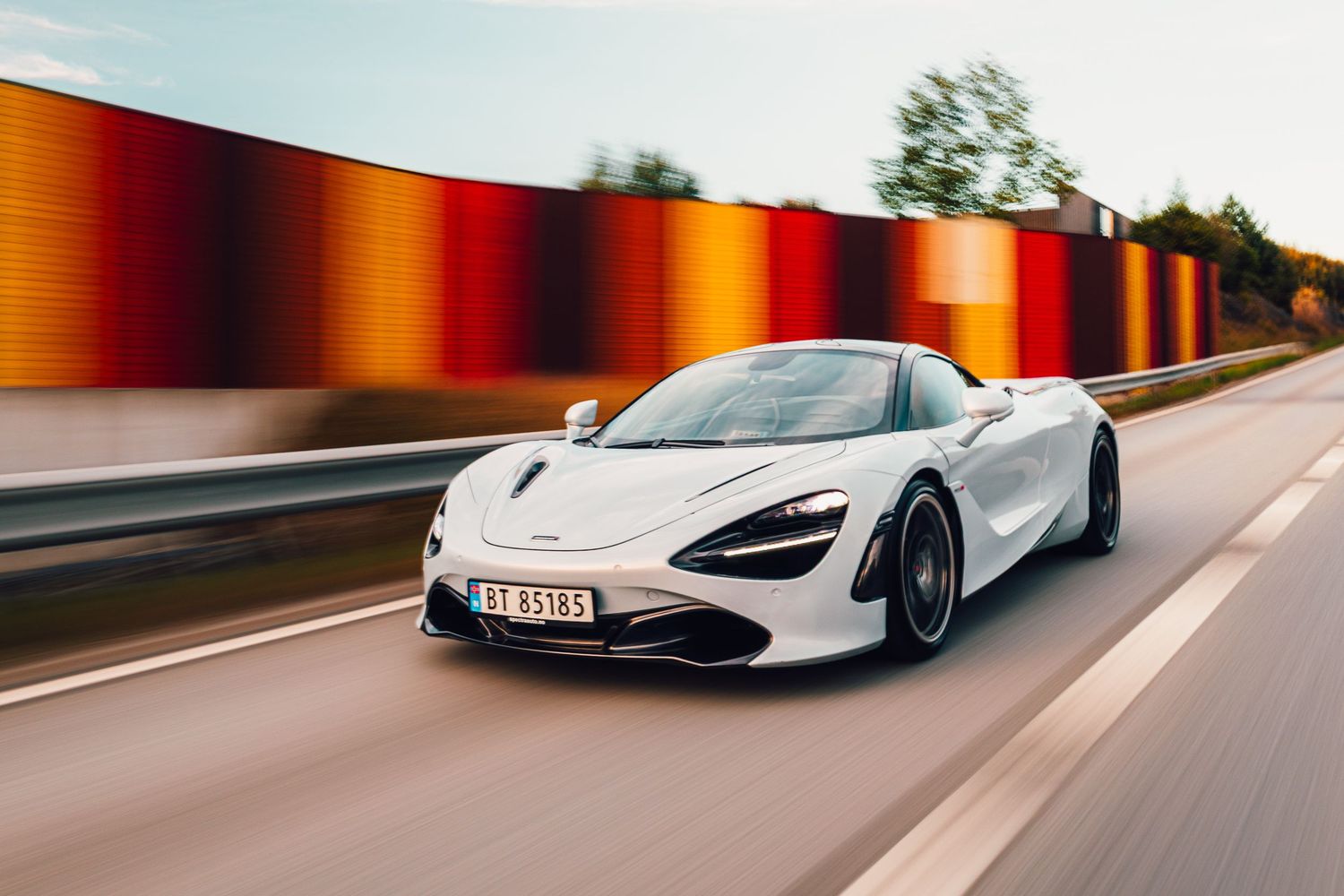 White McLaren 720S driving on road with colorful striped barrier wall.