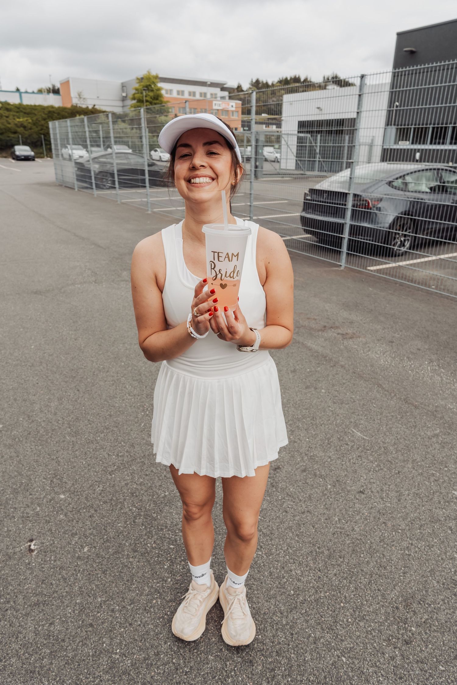 Person in white tennis outfit holding a drink in a parking lot.