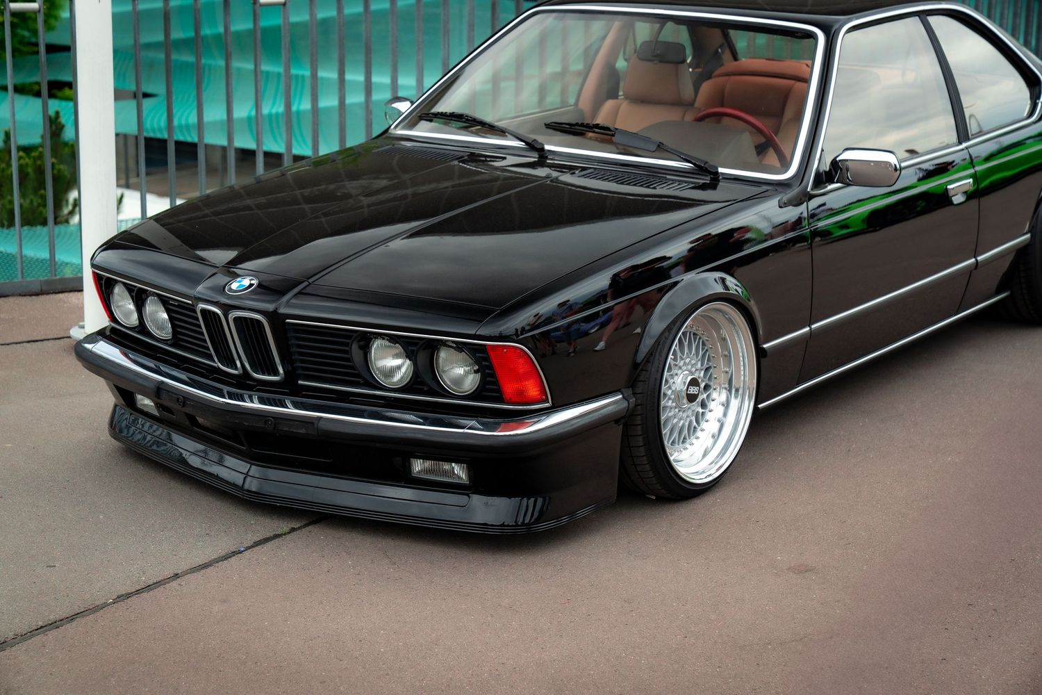 A black BMW E24 6-series with lowered suspension and silver BBS mesh wheels sits on a concrete surface.