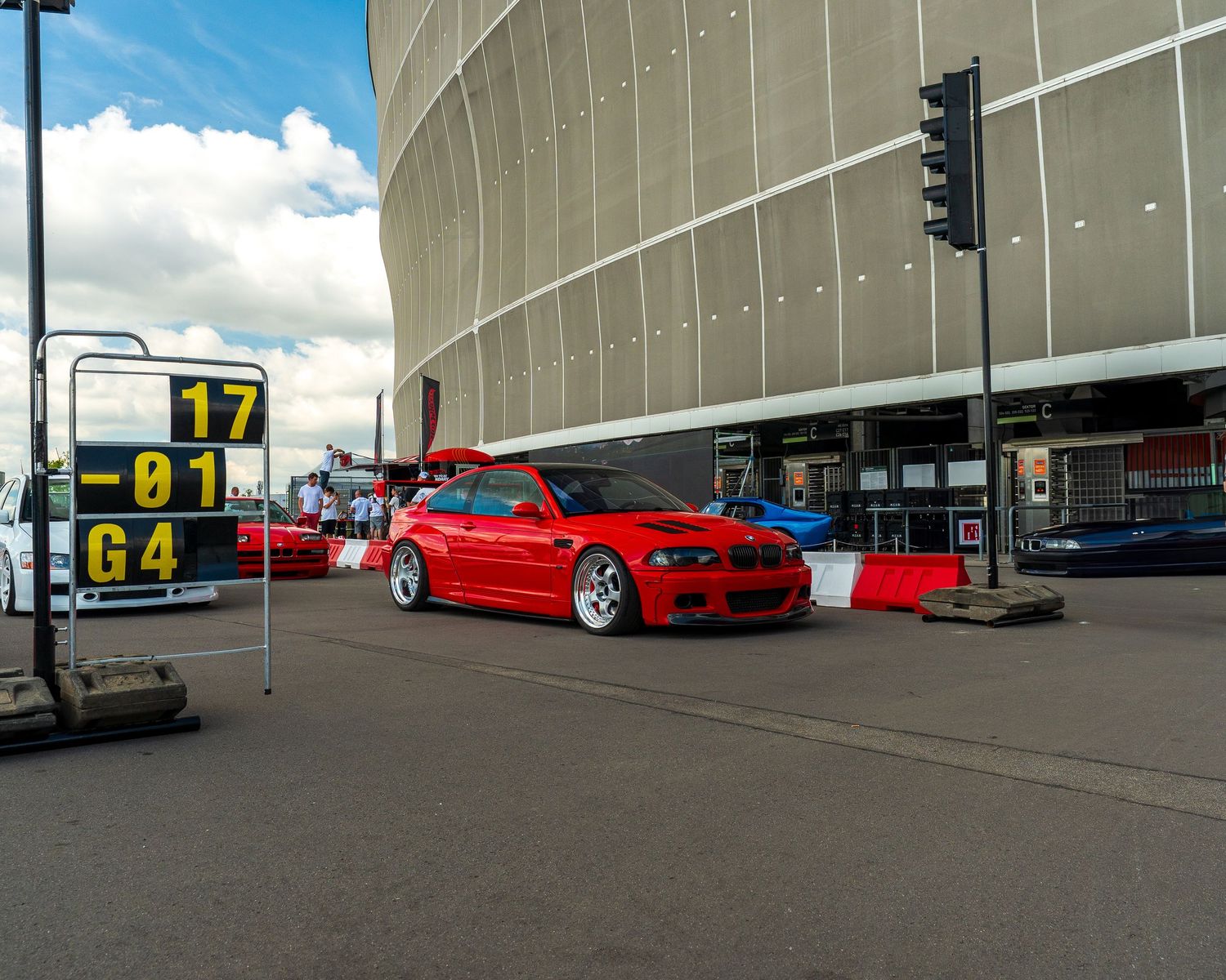 A red modified BMW M3 sports car parked outside a modern stadium building with traffic signals in view.