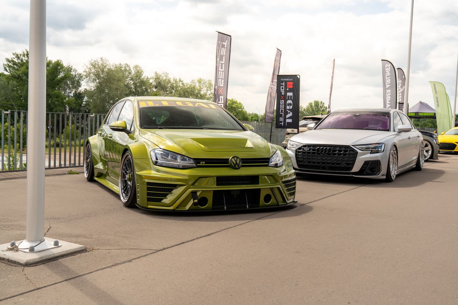 A lime green Volkswagen Golf and silver Audi RS3 displayed at an outdoor car show with banner flags.