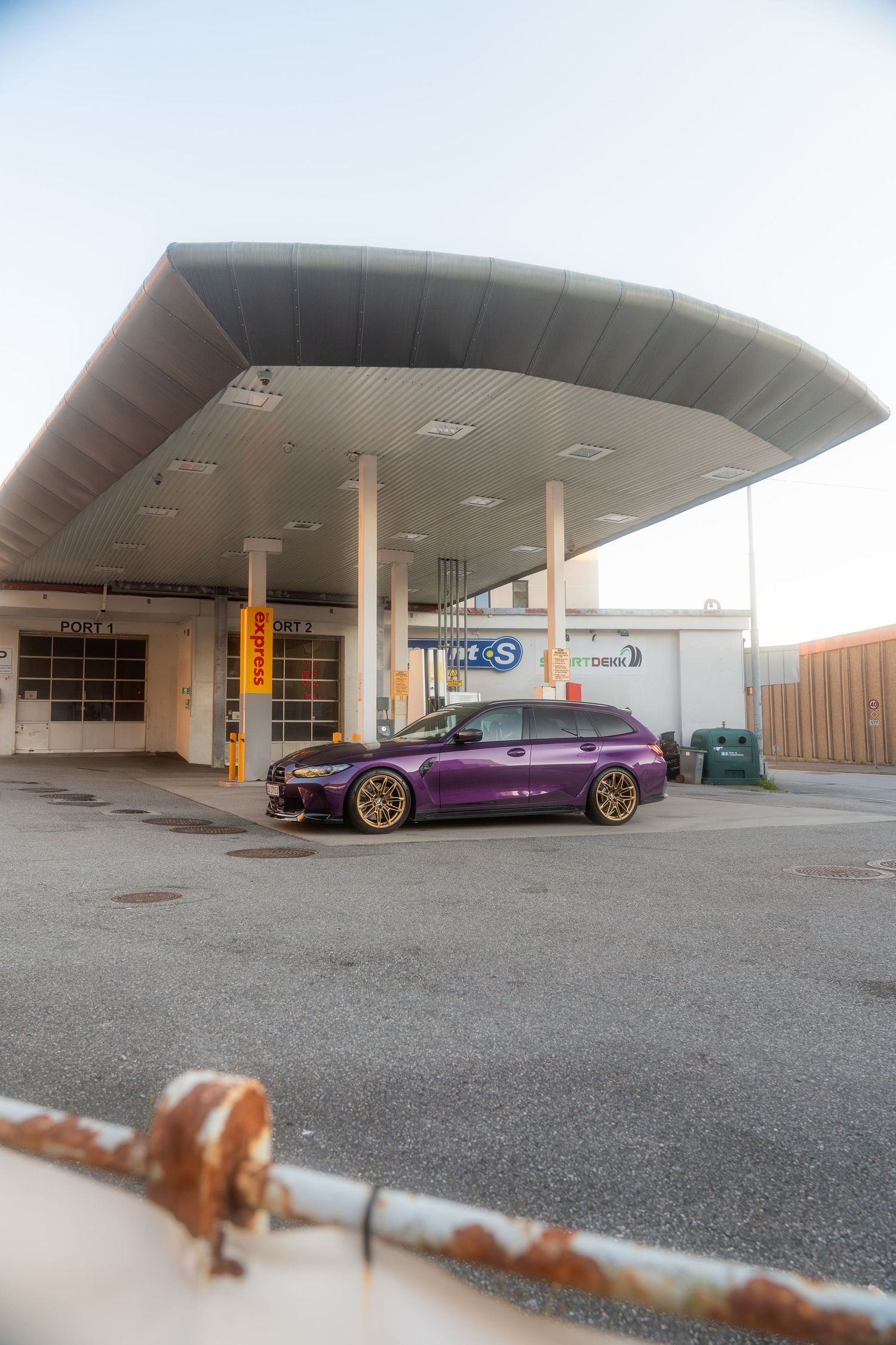 Purple BMW M4 parked underneath a modernist concrete canopy structure in an empty lot.