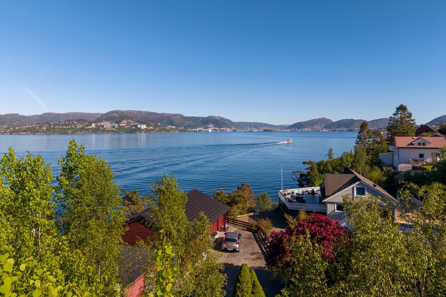 Scenic waterfront view of a calm blue fjord with lush green trees, red and white houses, and distant hills under a clear sky.