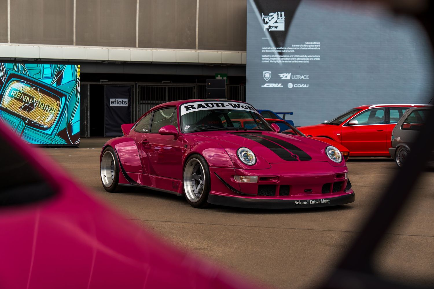 A modified pink Porsche 911 with wide body kit displayed at a car show.