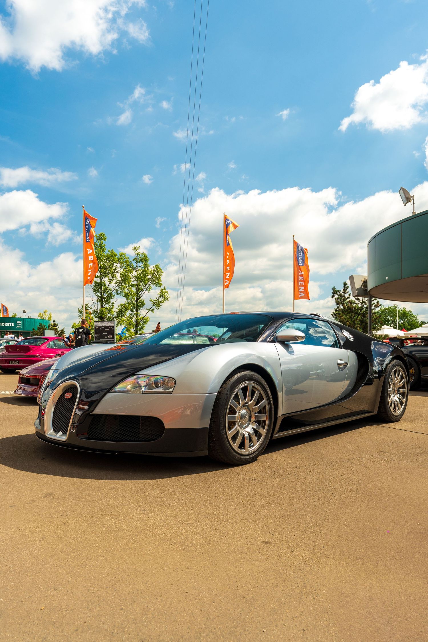 A silver and black Bugatti Veyron supercar parked outdoors with orange flags flying in the background under blue skies.