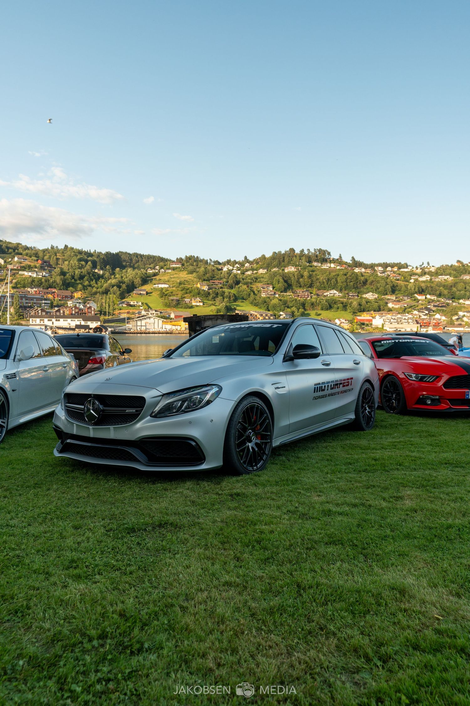 Sleek silver Mercedes-AMG vehicles lined up on grass with scenic waterfront and hillside views in the background.