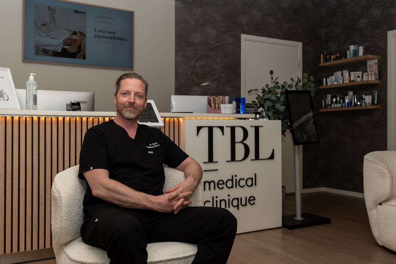Man in black scrubs sits in a cream armchair beside the TBL Medical Clinique reception desk in a modern, wood-paneled clinic lobby.