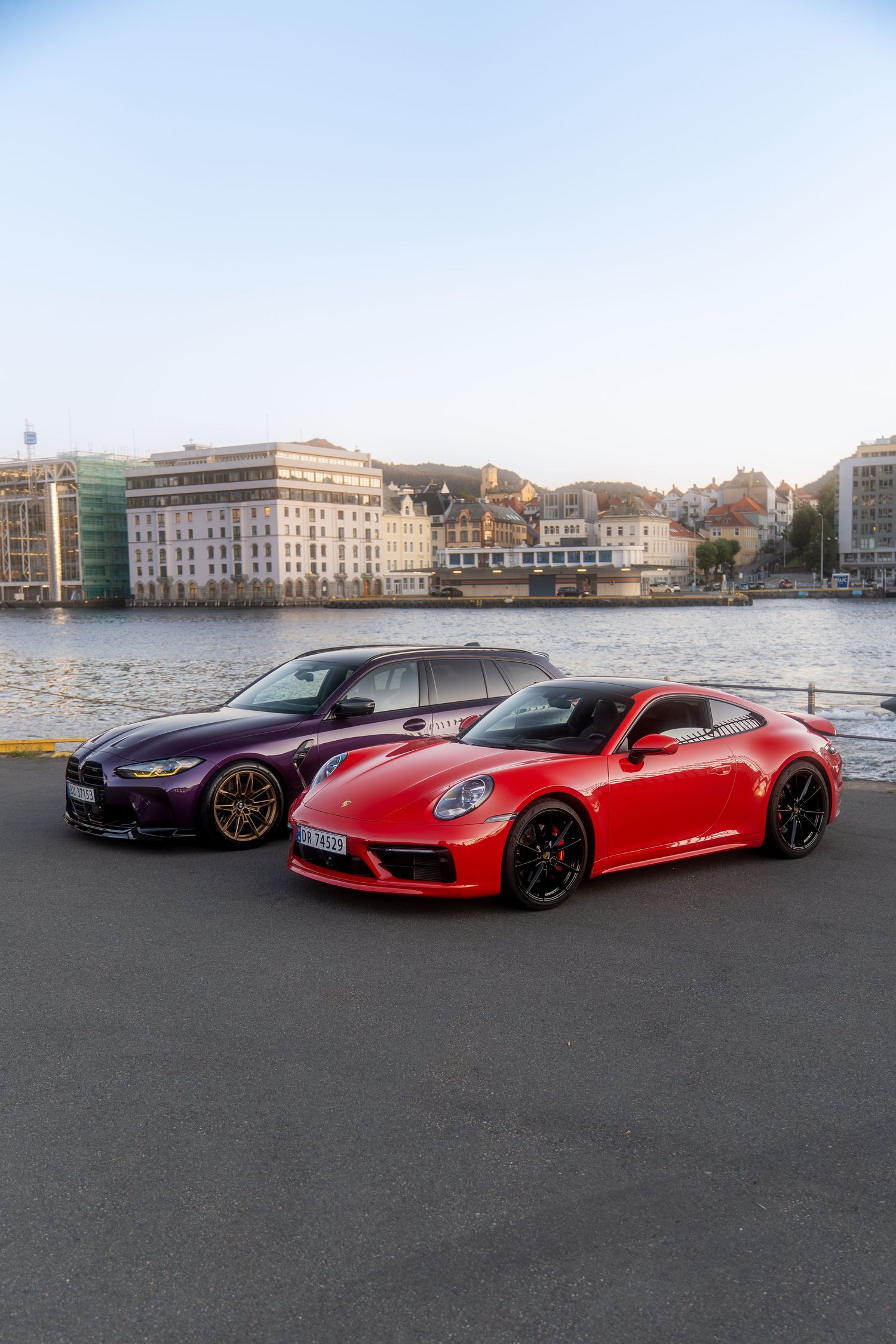 Collection of red and purple luxury sports cars lined up against a coastal cityscape backdrop.