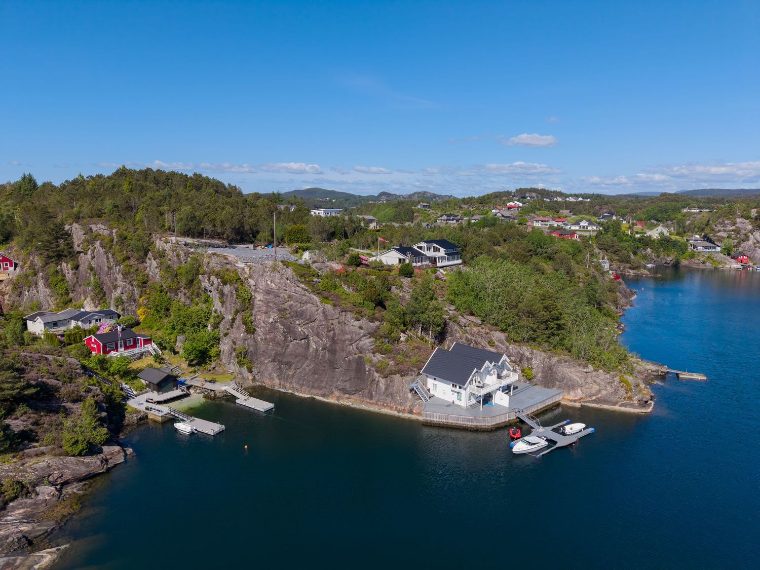 Aerial view of a white waterfront house with a private dock and boats on a calm blue fjord, surrounded by rocky cliffs and green forest.