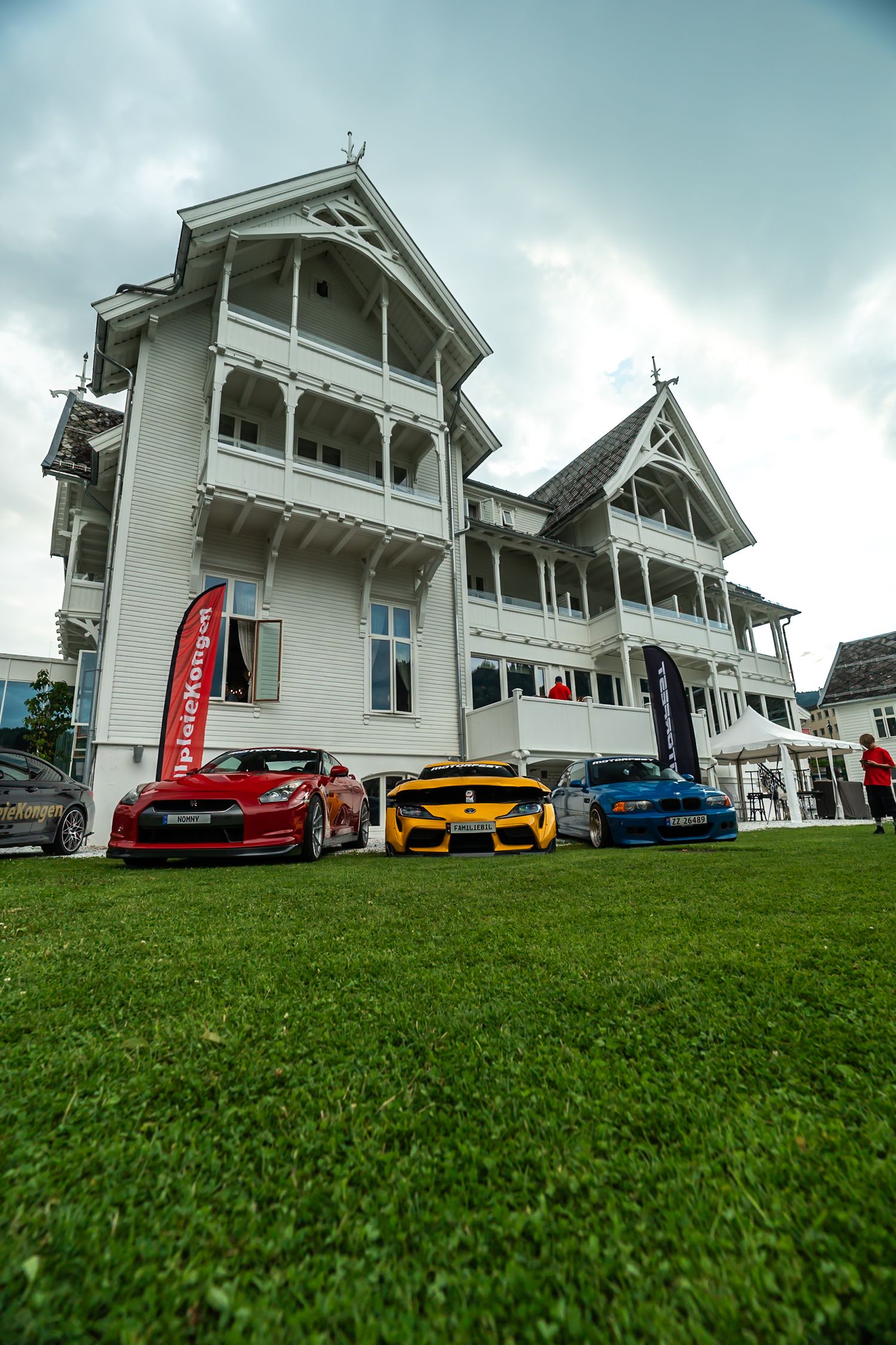 Red, yellow and blue sports cars parked on a green lawn in front of a white historic wooden villa with ornate balconies under a cloudy sky.