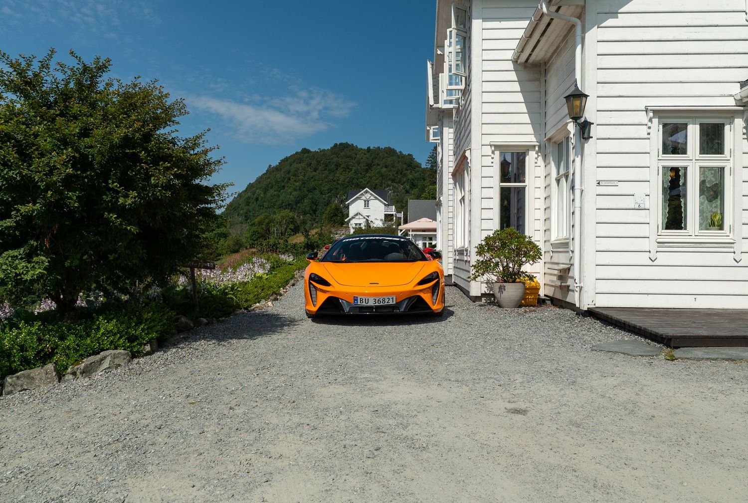 Orange McLaren sports car parked on gravel driveway beside white wooden house with mountain backdrop on sunny day.