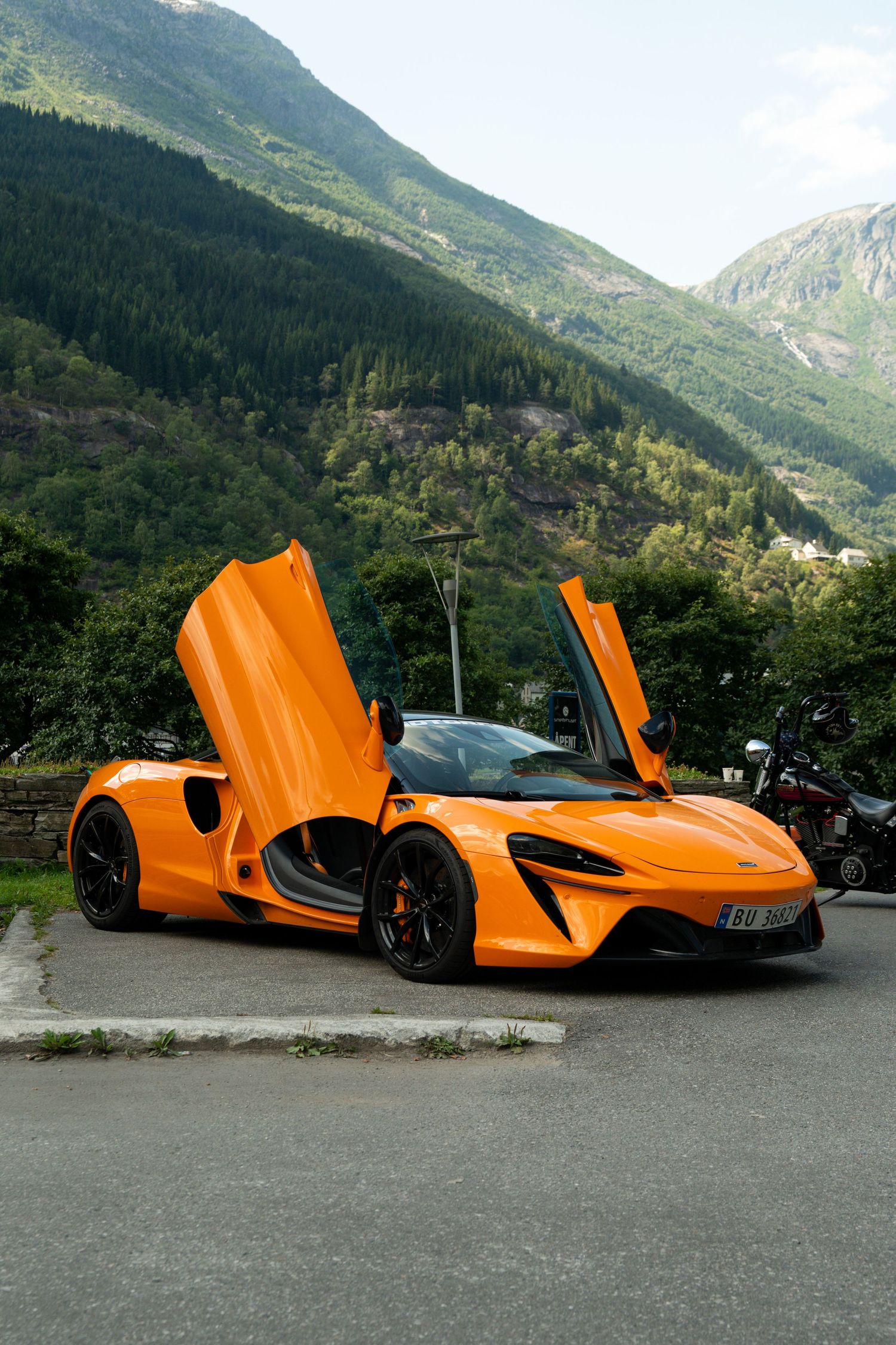 Orange McLaren supercar with dihedral doors open, parked on mountain road surrounded by lush green hills.