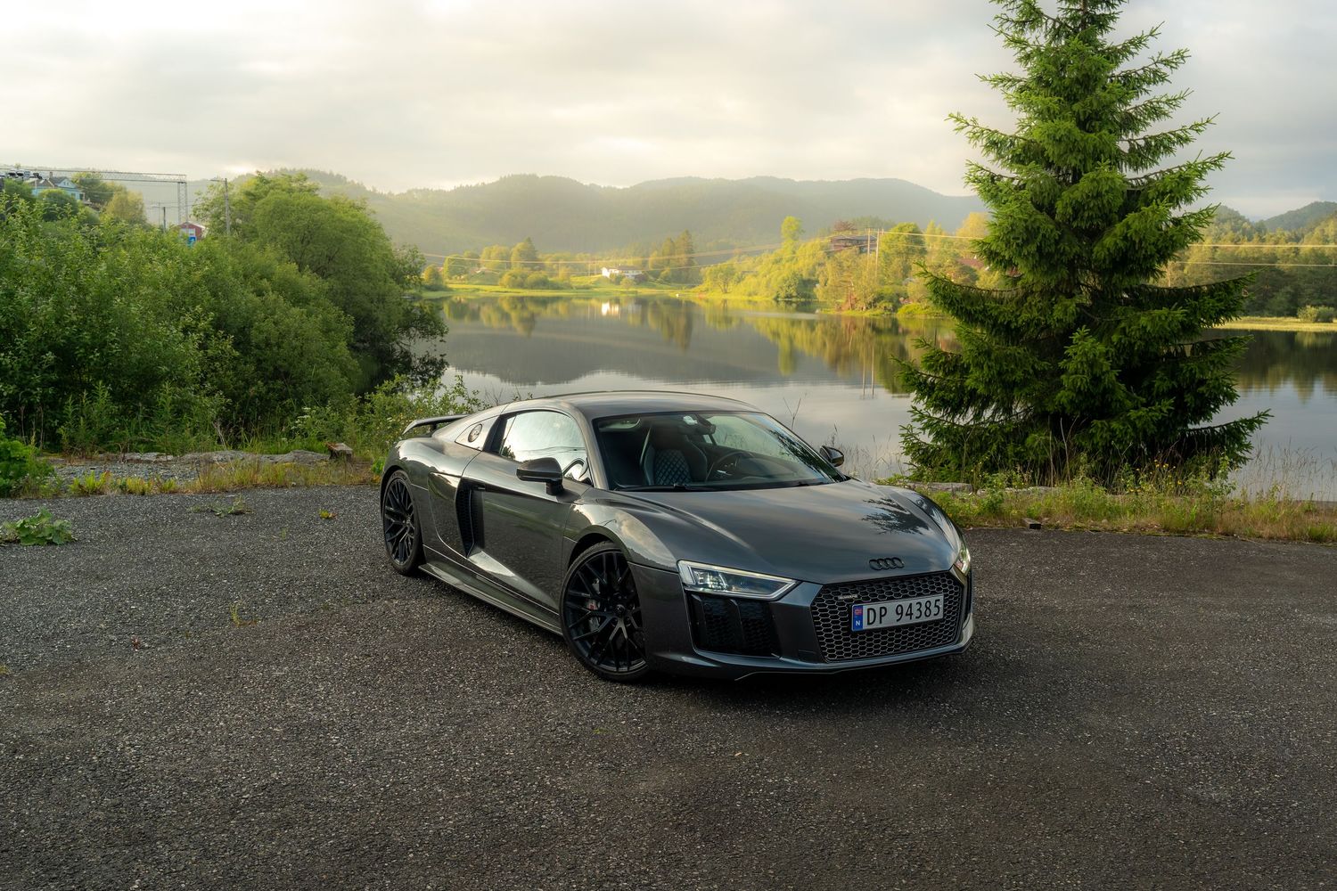 Black Audi R8 supercar parked at scenic lakeside location with mountains and trees in background.