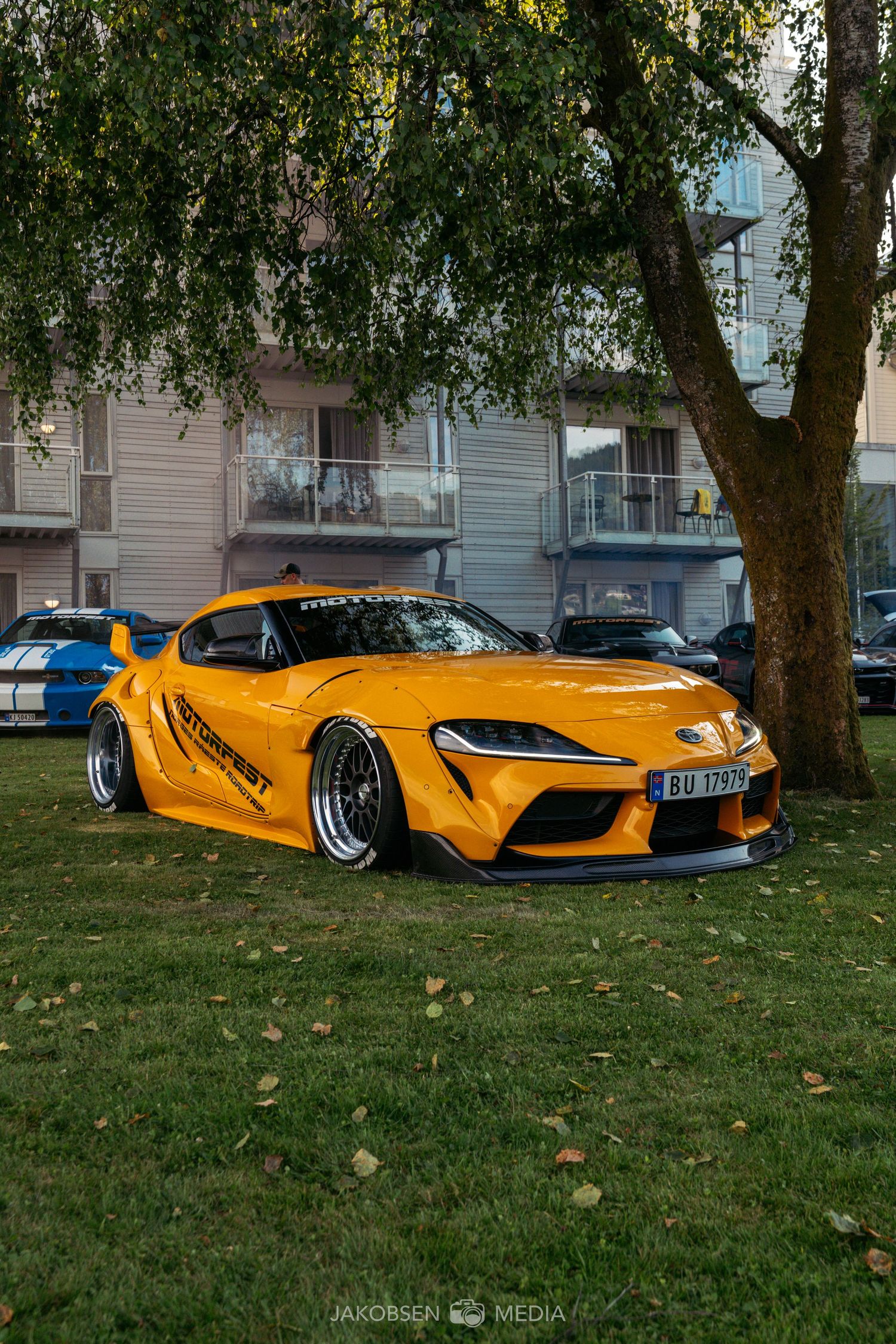 A McLaren sports car in striking yellow color resting on grass beneath overhanging birch trees.
