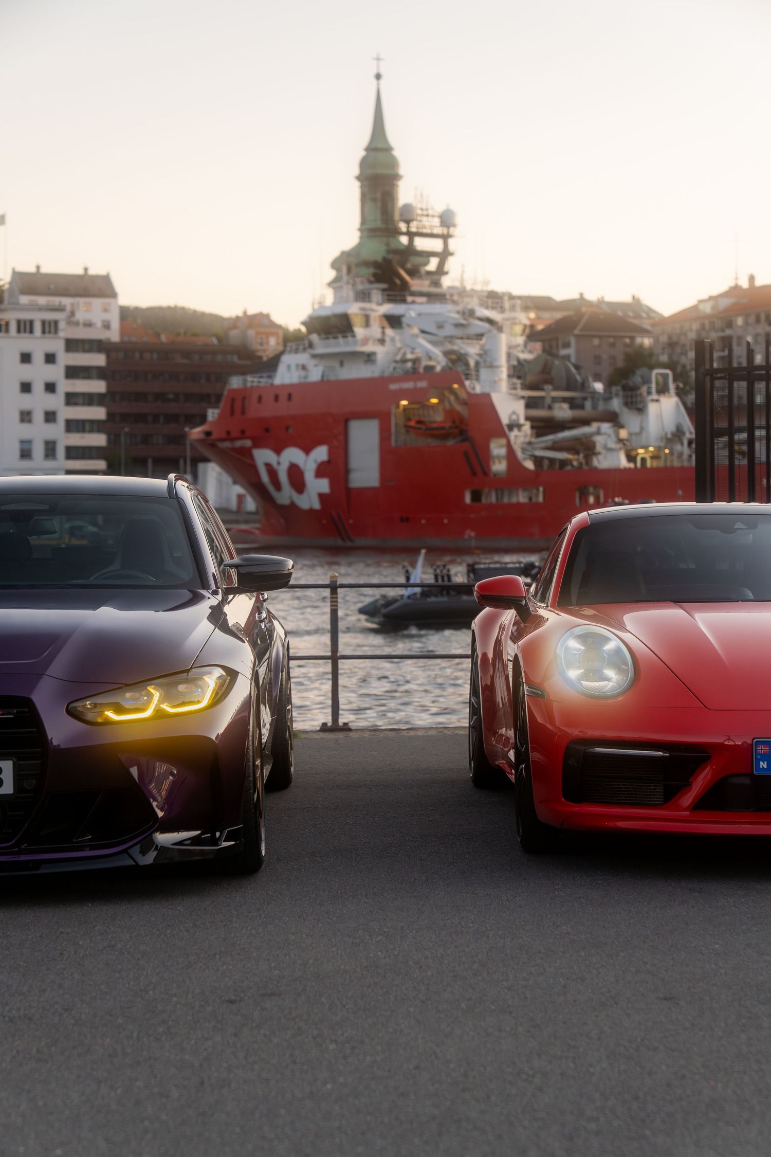 A row of luxury sports cars parked at sunset along a waterfront with boats and buildings in the background.