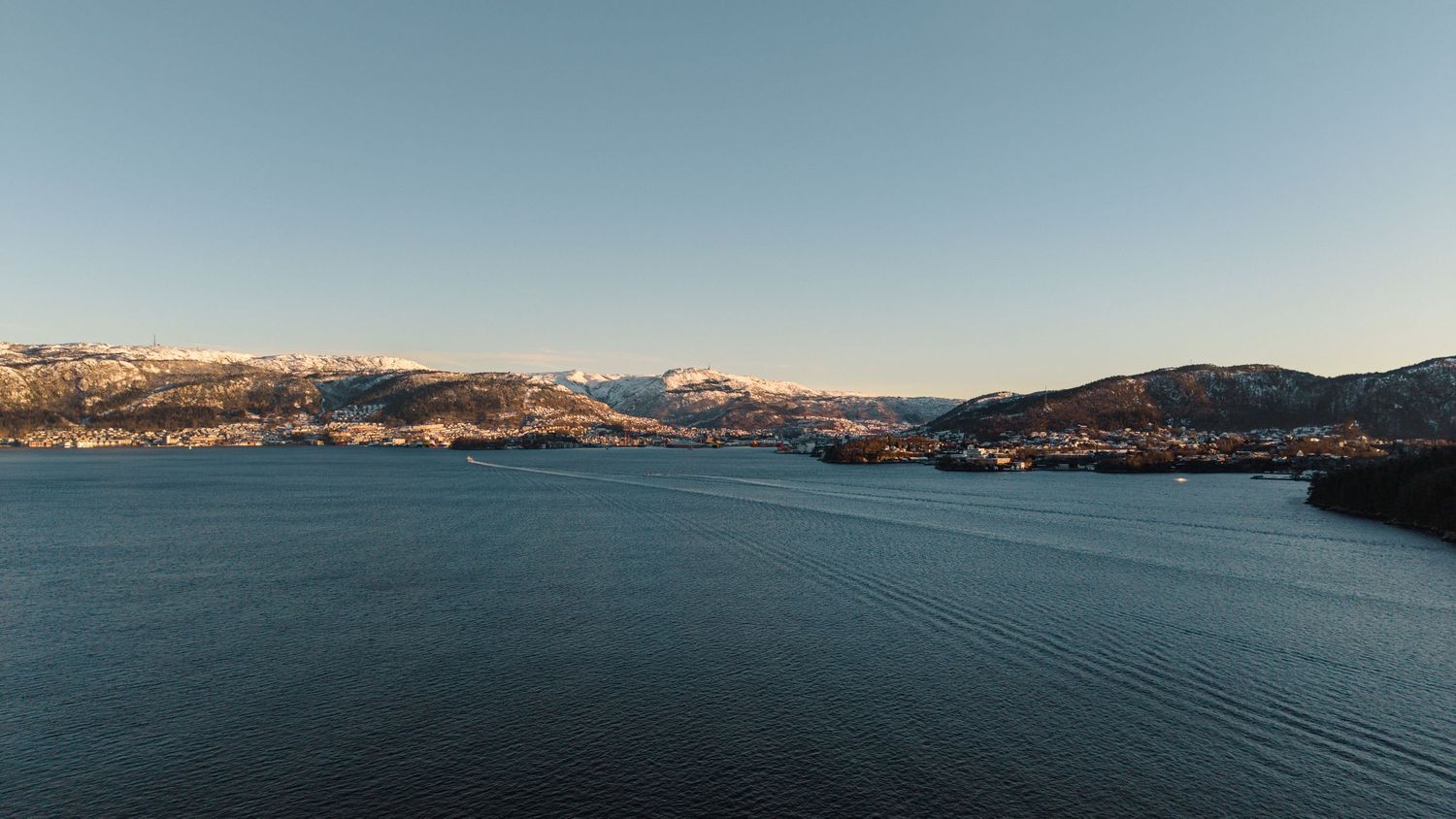 Aerial view of a calm fjord surrounded by snow-capped mountains and a small town under a clear blue sky.