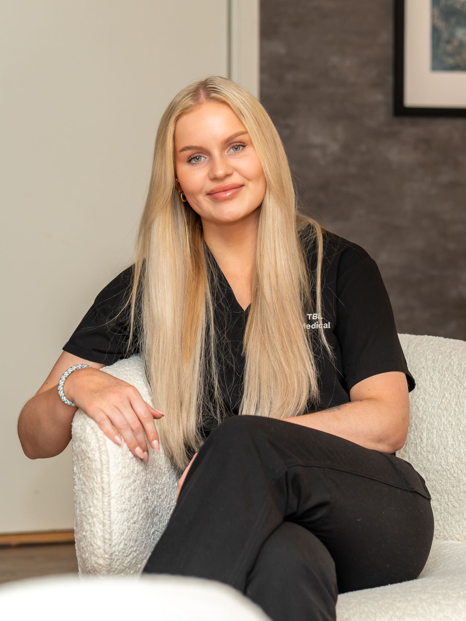 Smiling woman with long blonde hair in black medical scrubs sits relaxed on a cream textured armchair in a modern, softly lit clinic.