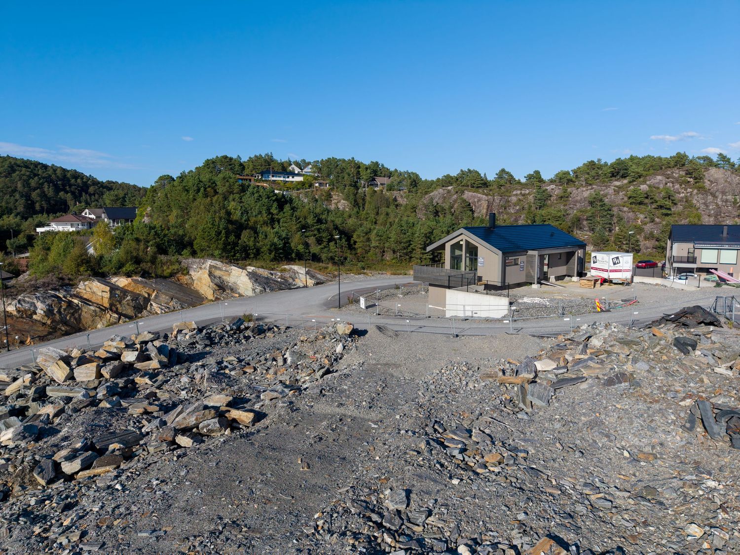 Modern cabin with a blue roof on a rocky hillside plot, surrounded by forested hills and a clear blue sky, with a vehicle parked nearby.
