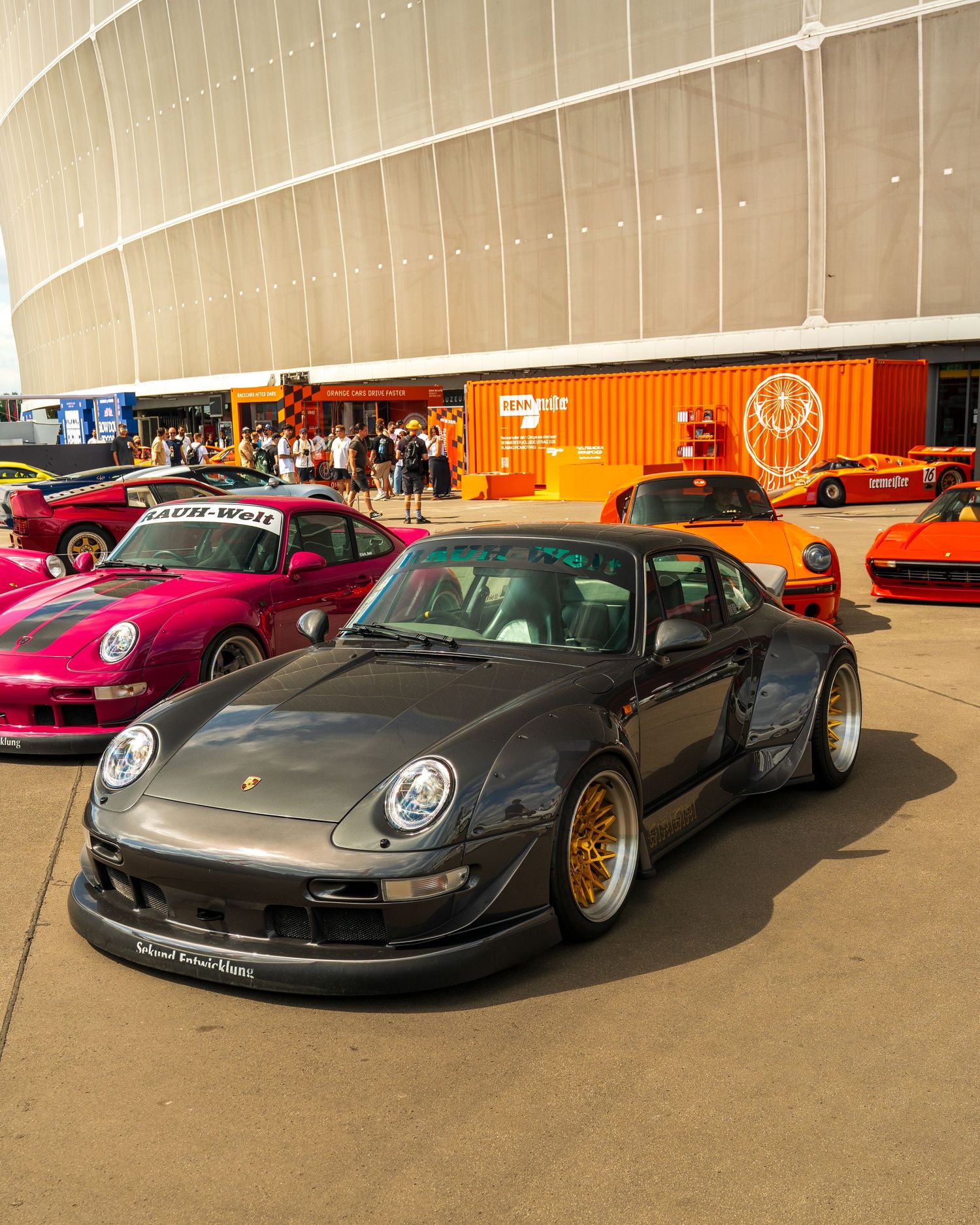Modified gray Porsche 911 with gold wheels parked at an indoor car show next to other sports cars.