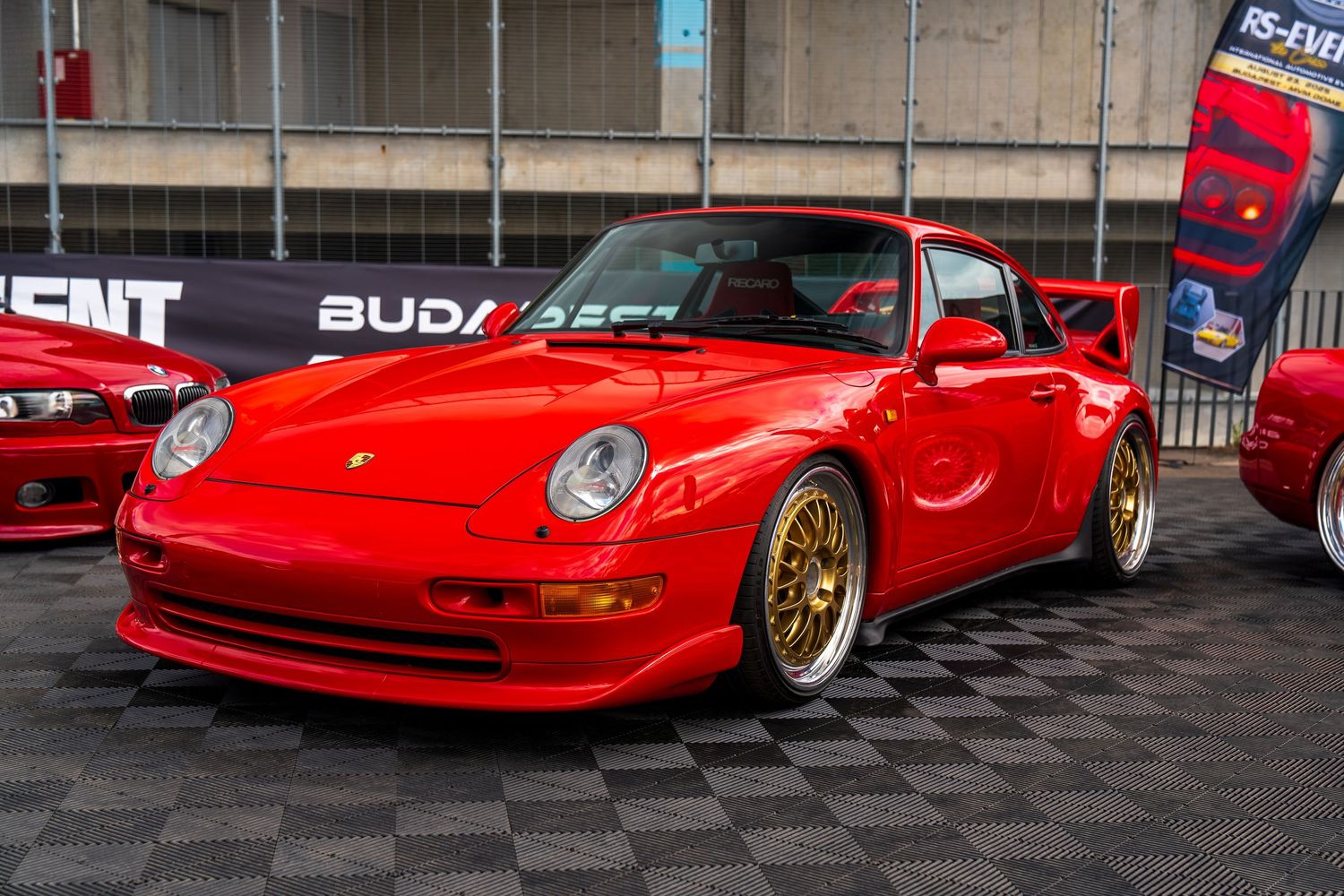 Red Porsche 993 with gold wheels displayed on checkered flooring at an auto show.
