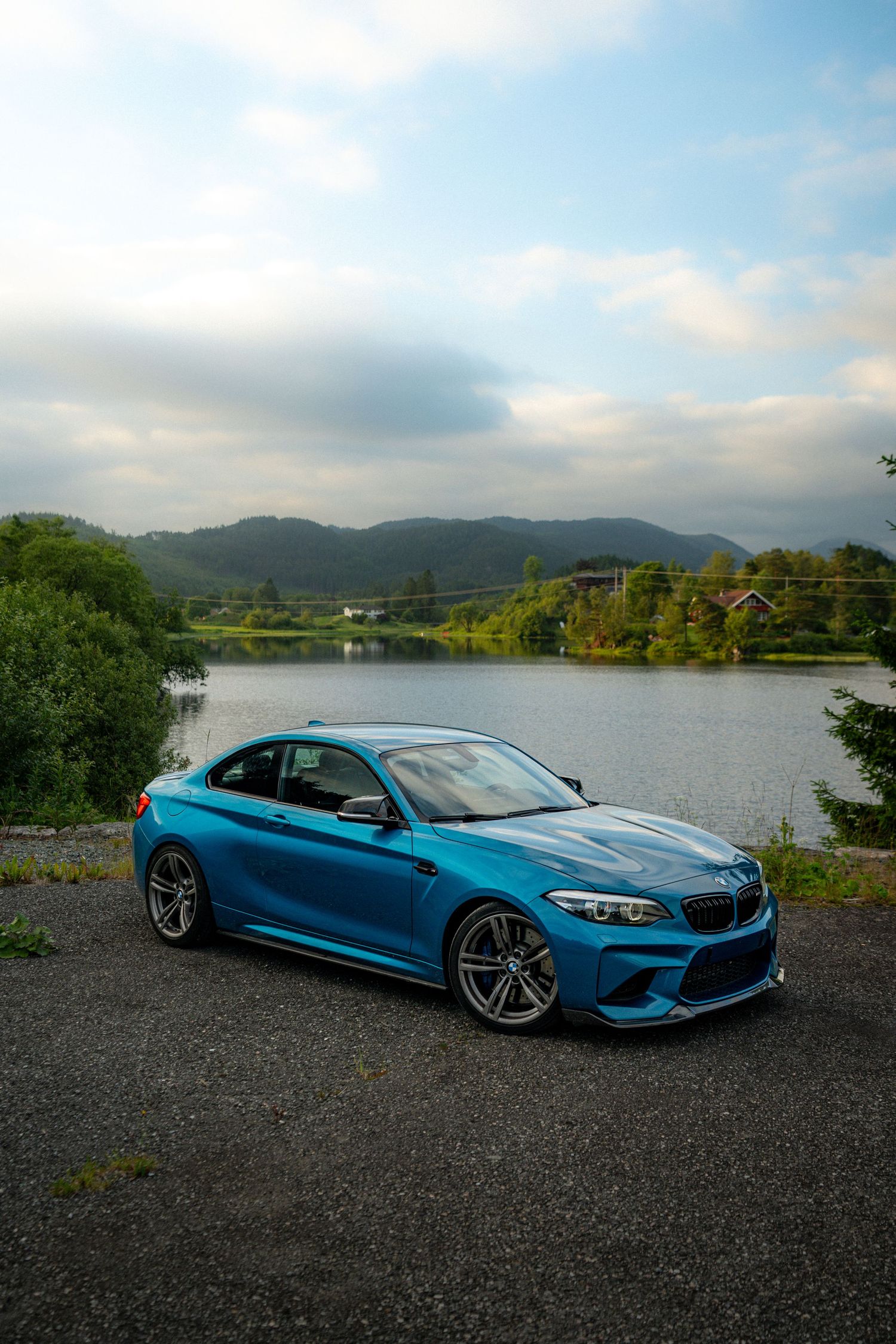 Modified blue BMW M2 cars parked by a lake with mountains in the background.