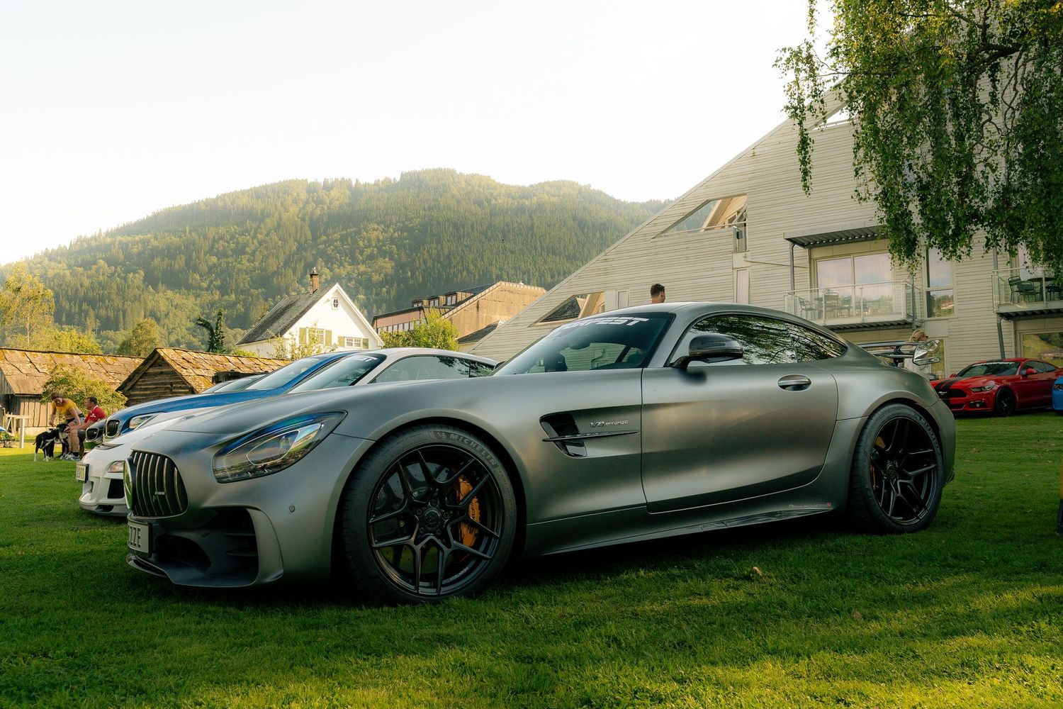 Silver Mercedes-AMG GT R sports car with black alloy wheels parked on a green lawn near alpine-style buildings and forested hills.