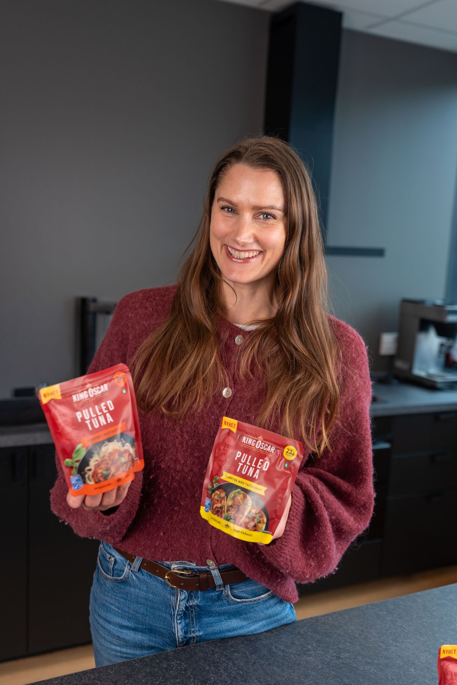 Smiling woman in a burgundy sweater holds two pouches of Pulled Tuna in a modern kitchen with dark cabinets.