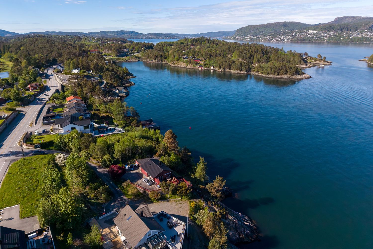 Aerial view of a calm blue fjord with forested peninsulas, scattered waterfront houses and a winding coastal road under a clear sky.