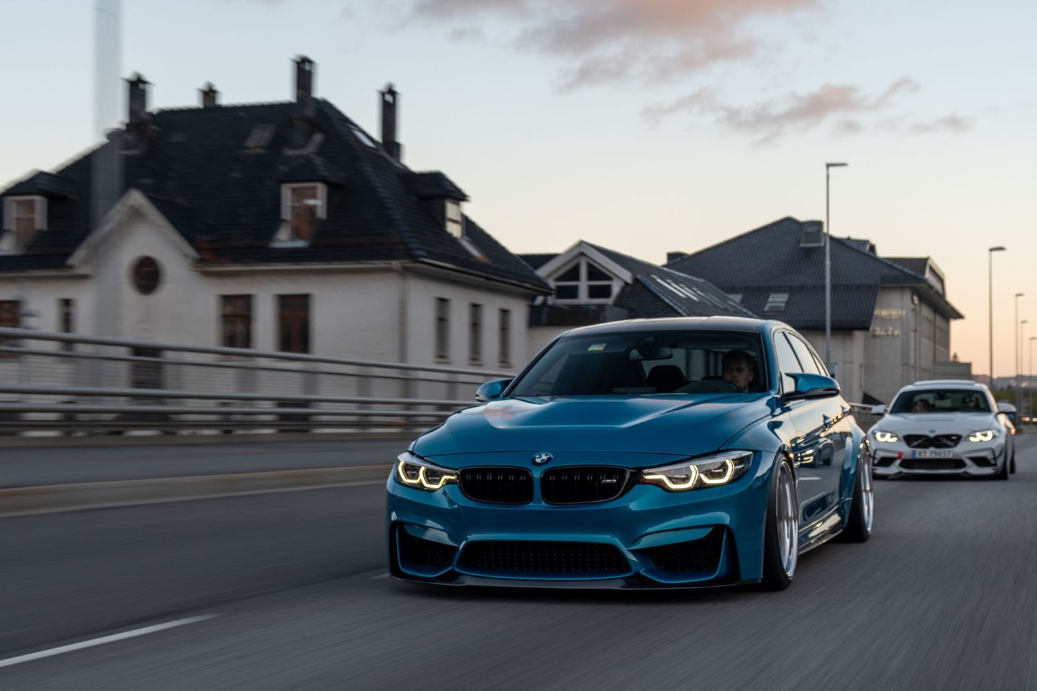Lowered blue BMW M3 driving on urban street at sunset with residential buildings.