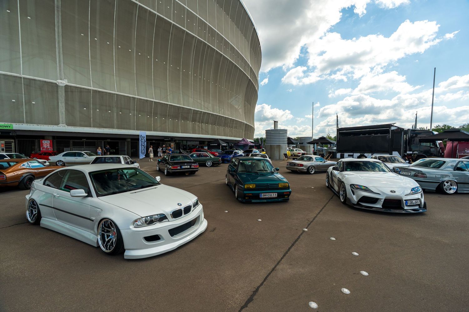 Modified cars parked outside a large stadium building during a car show event.