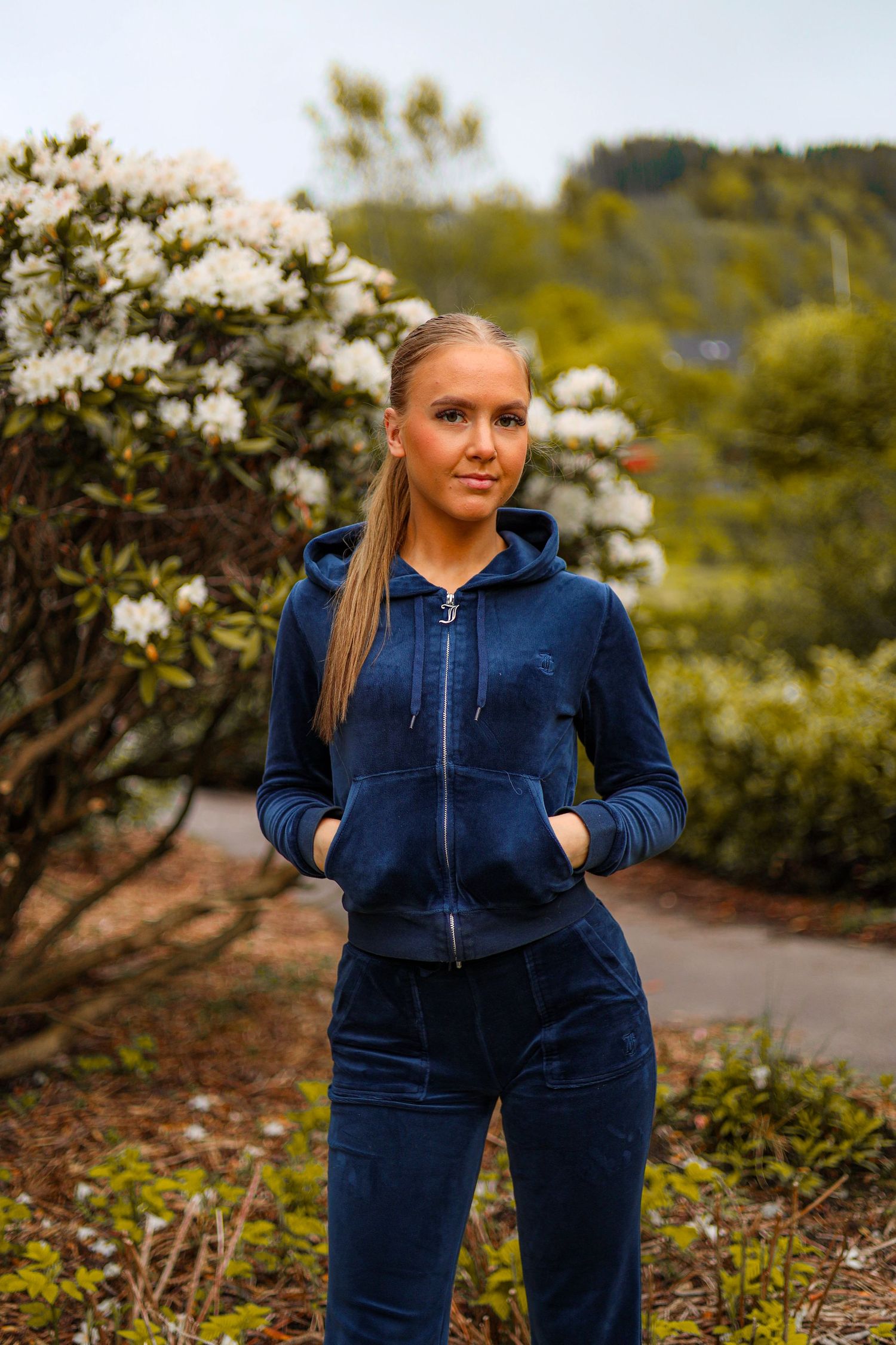 A person in a navy blue velvet tracksuit stands on a garden path with white flowering bushes in the background.