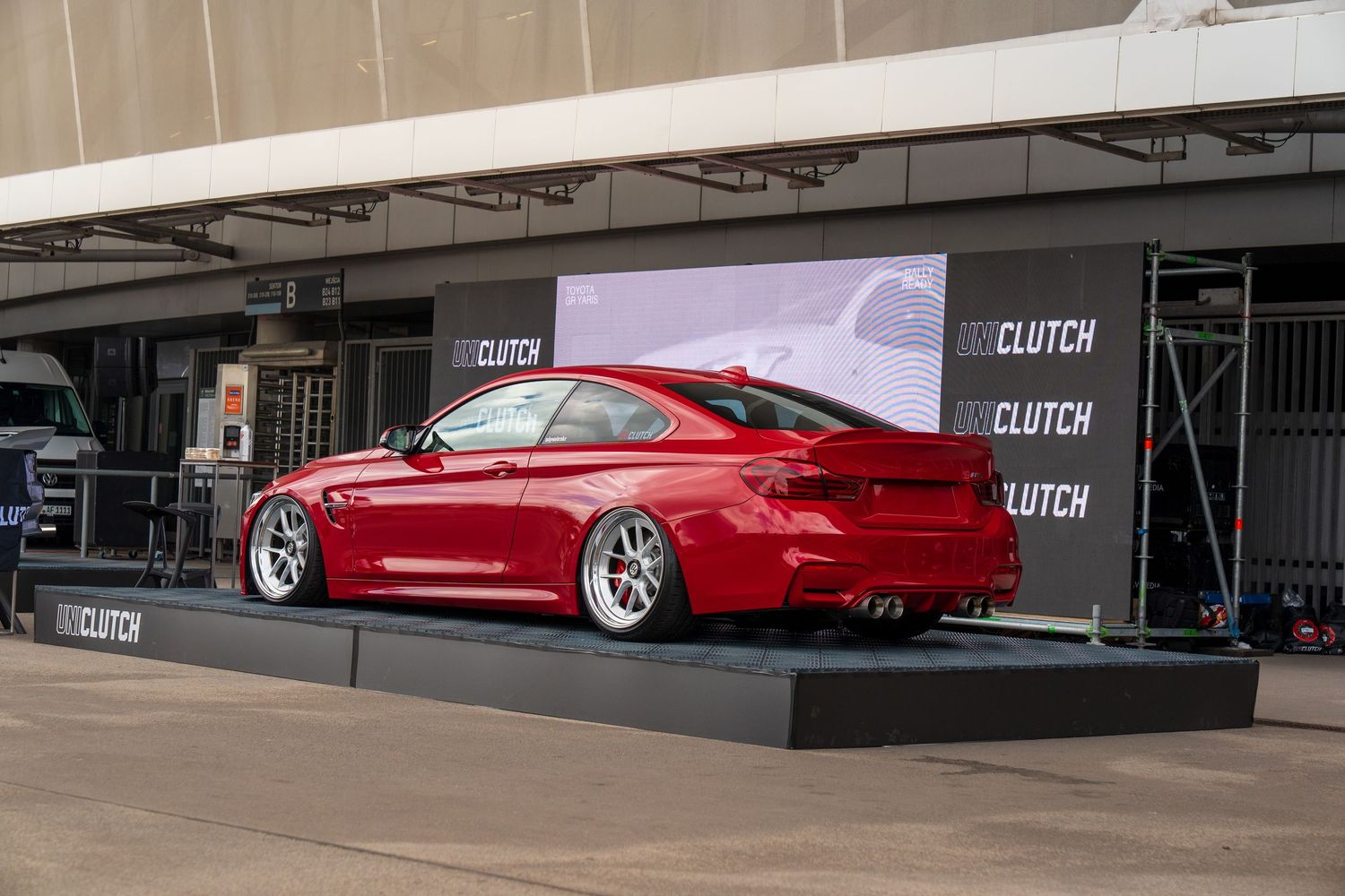 A lowered red BMW M4 on display at an event with promotional backdrop and Clutch branding.