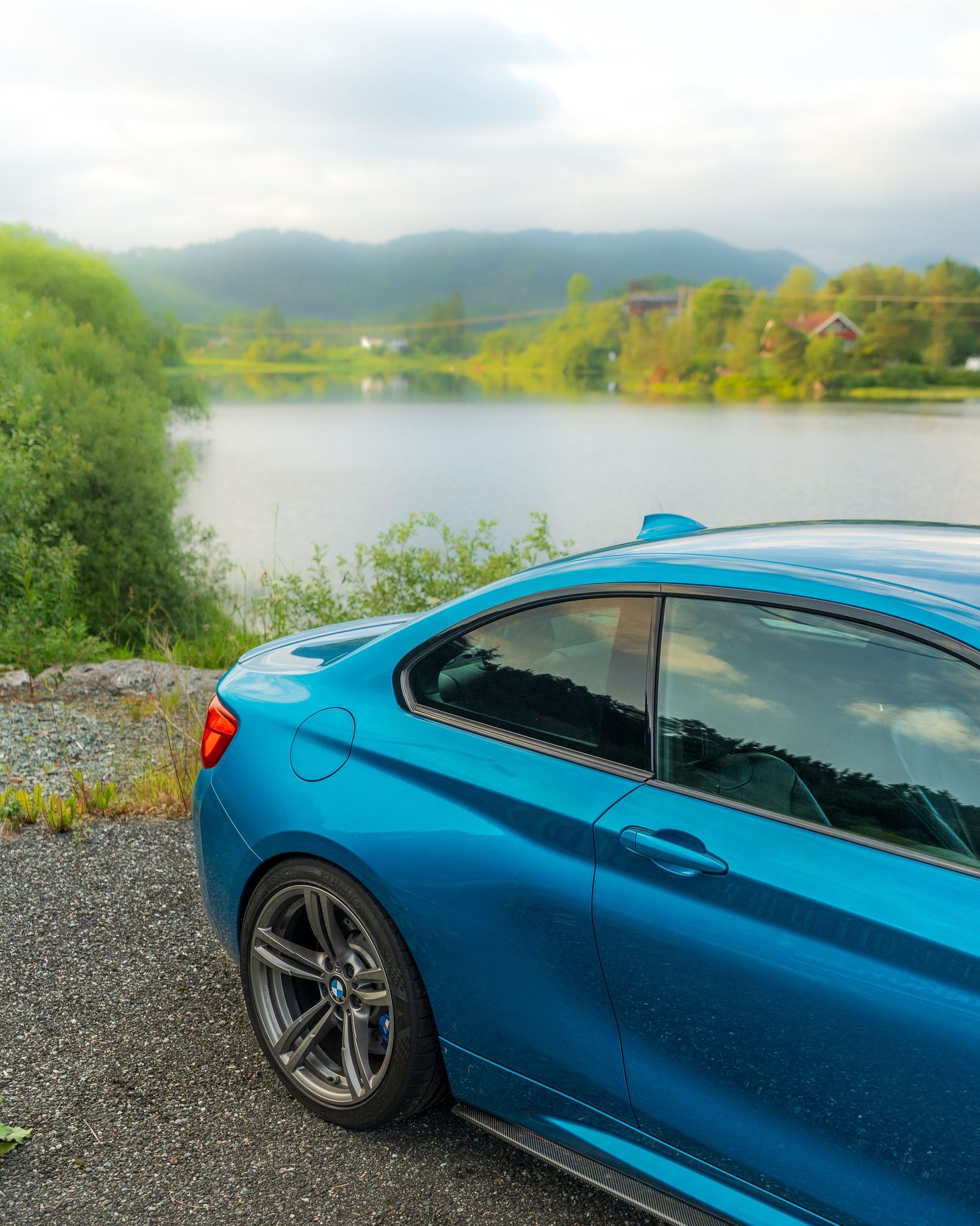 Side profile of a blue BMW M2 parked at scenic overlook with lake and mountains in background.