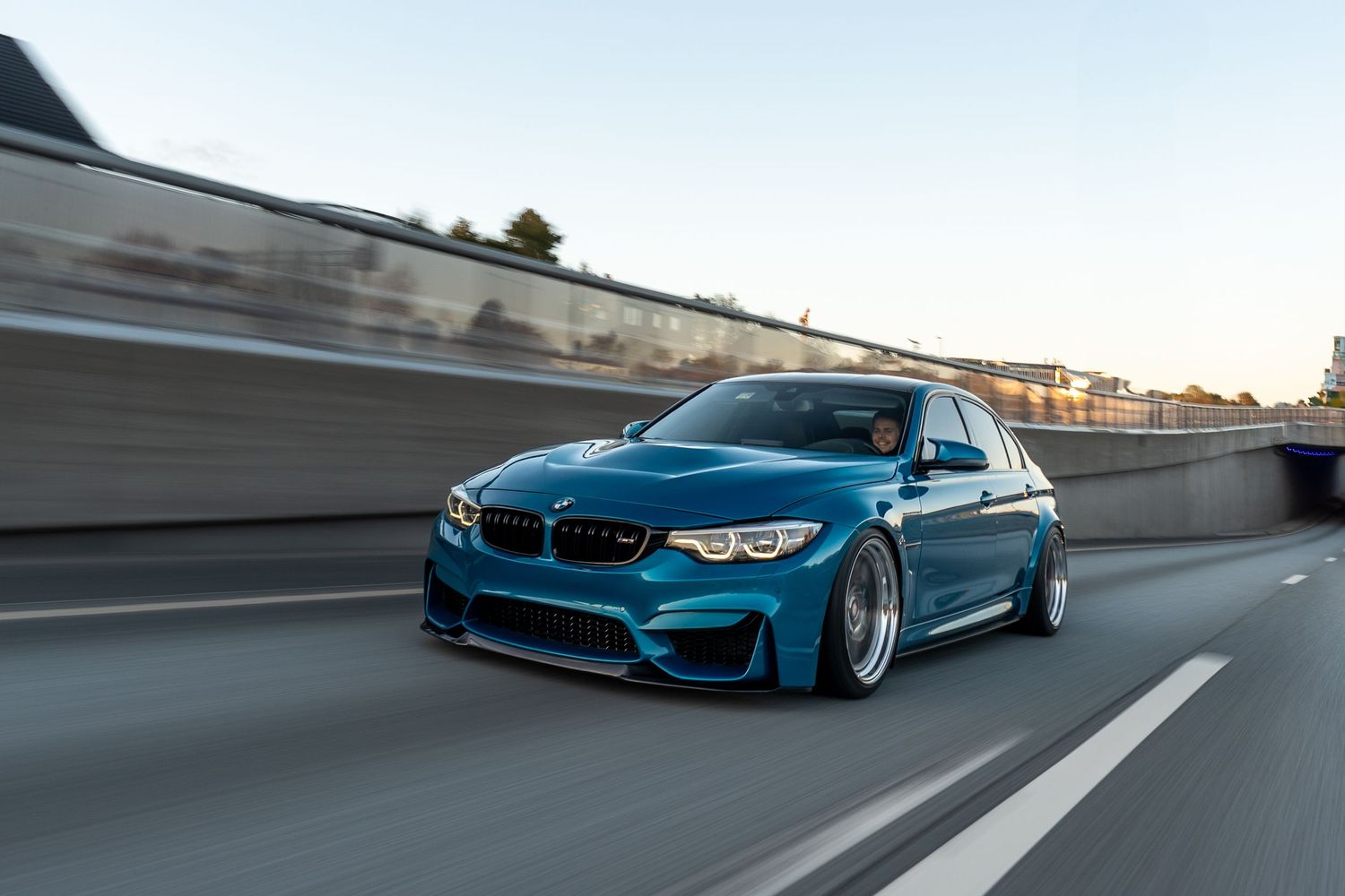 Rolling shot of blue BMW M3 on highway showing modified stance and custom wheels.