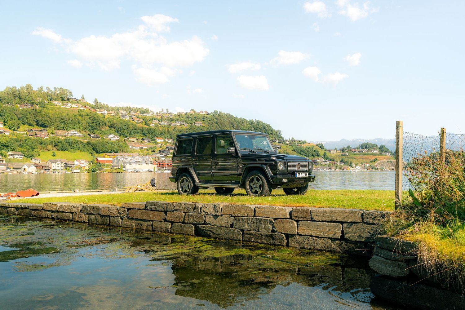 Black Mercedes-Benz G-Class SUV parked on a grassy lakeside lawn behind a stone wall, with a scenic village and forested hills beyond.