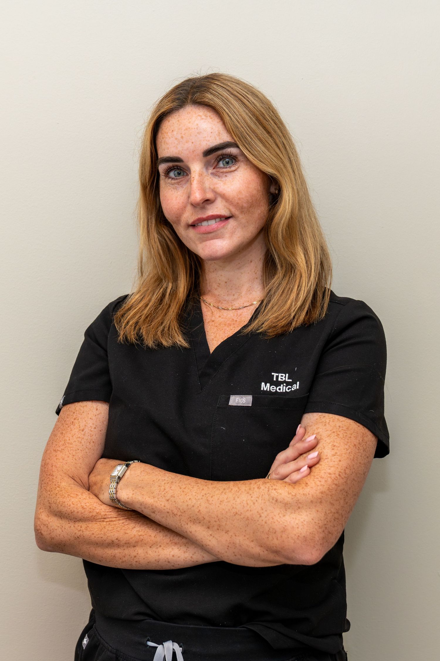 Smiling woman with shoulder-length blonde hair wearing black TBL Medical scrubs and standing with her arms crossed against a neutral wall.