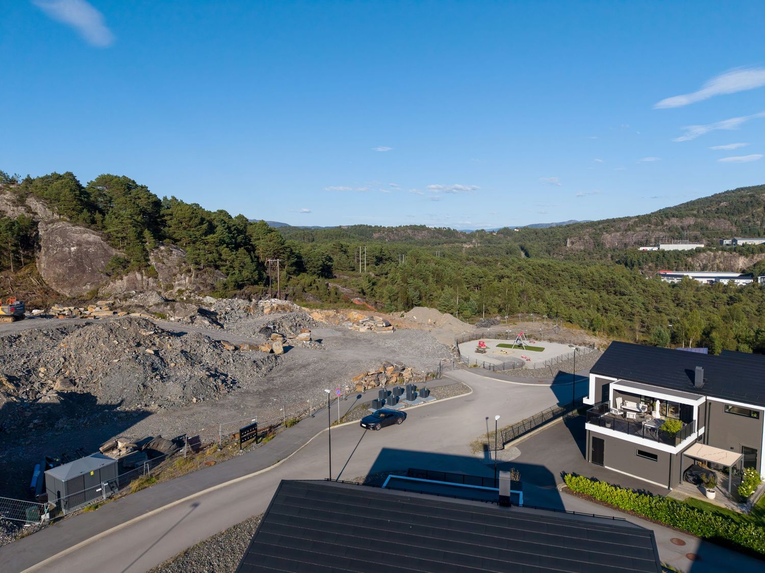 Aerial view of a hillside construction site with exposed rock and graded earth beside a paved road, modern homes, and forested hills.