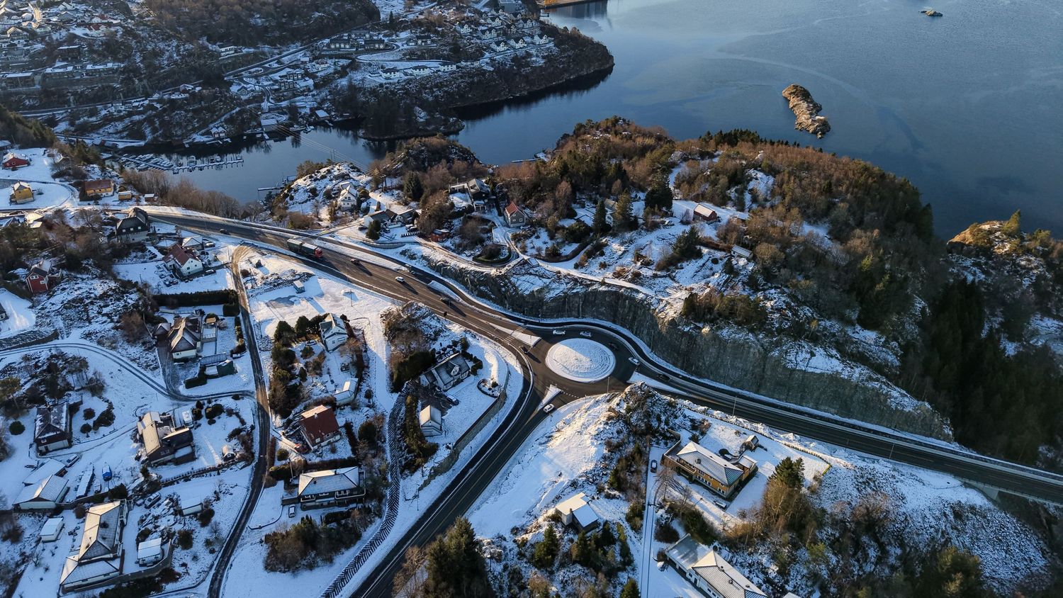 Aerial winter view of a snowy Scandinavian coastal town with a roundabout road, colorful houses, and icy blue water.