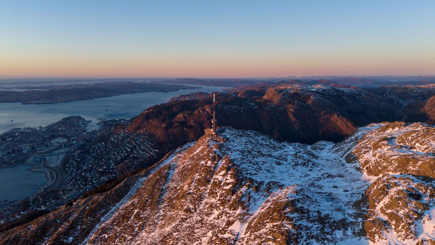 Aerial winter sunset view of snowy mountain peak with tower overlooking fjord and city in Norway.