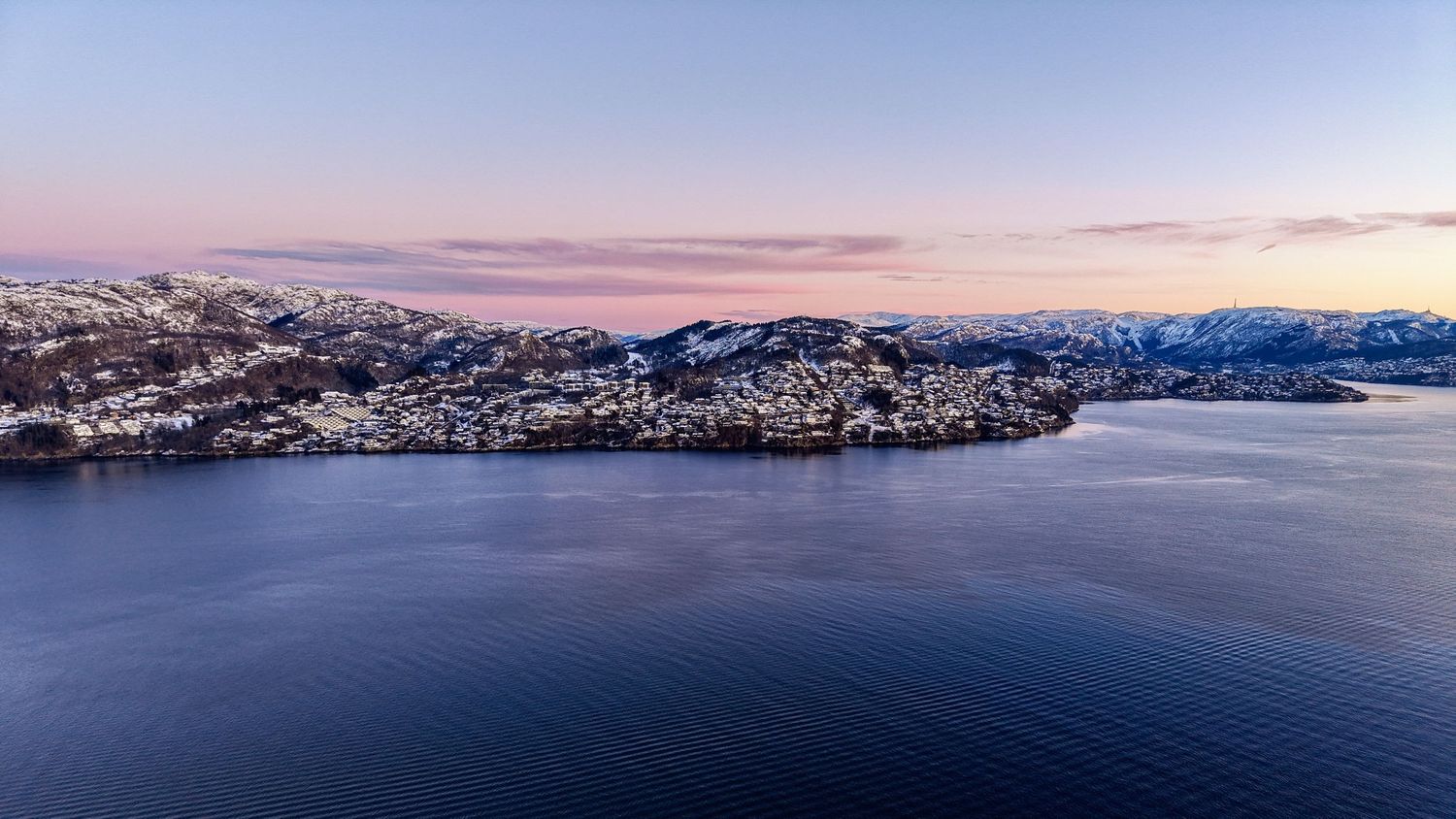 Aerial winter dusk view of a snowy coastal town reflecting pink skies over calm blue fjord waters in Norway.
