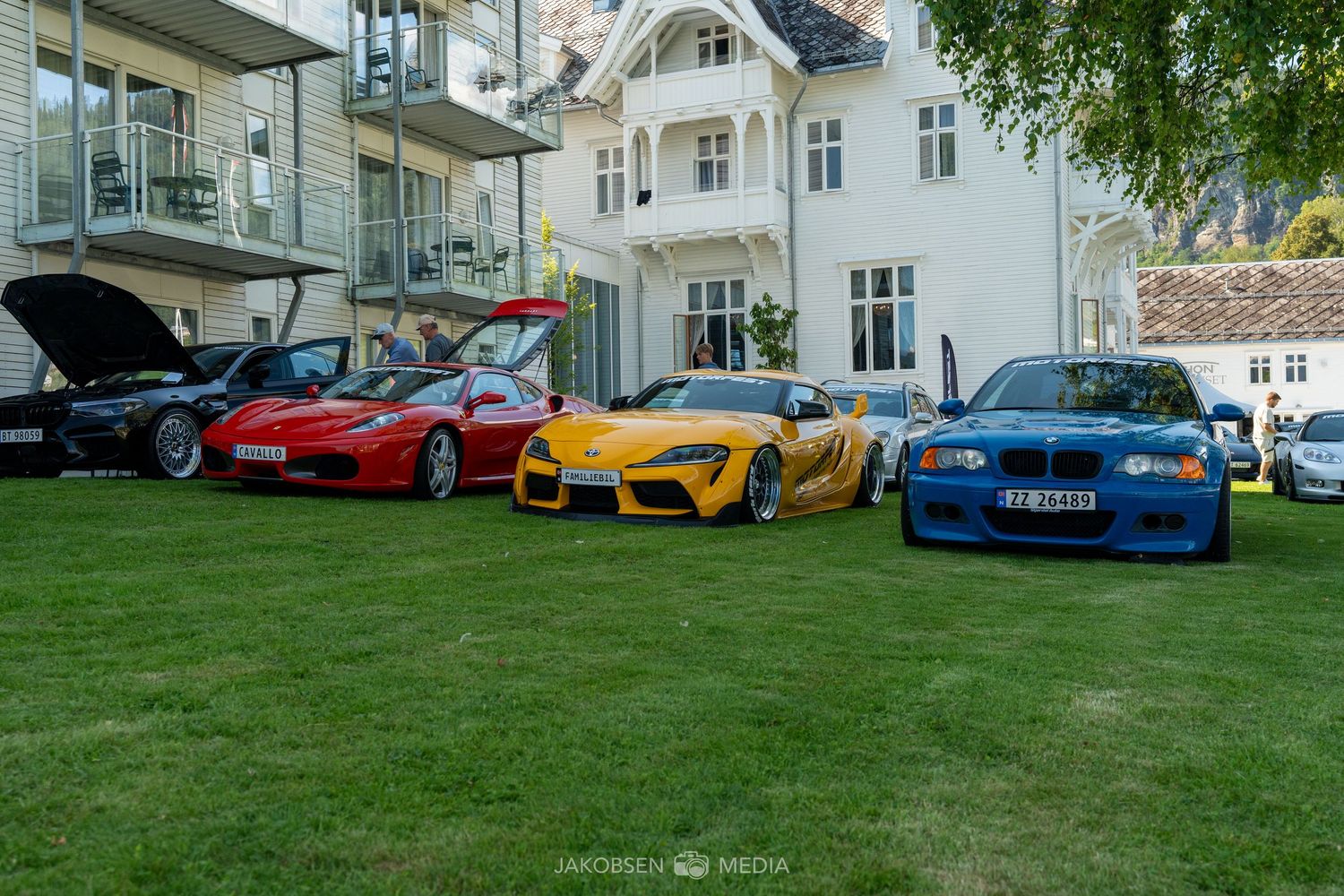Lineup of colorful sports cars including Ferrari and Porsche models displayed on grass in front of a white mansion.