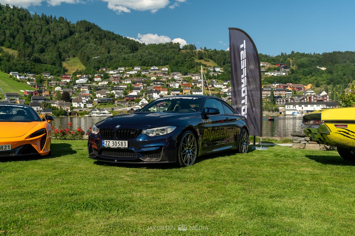 Luxury sports cars lined up at a scenic outdoor car show with mountains and greenery in the background.