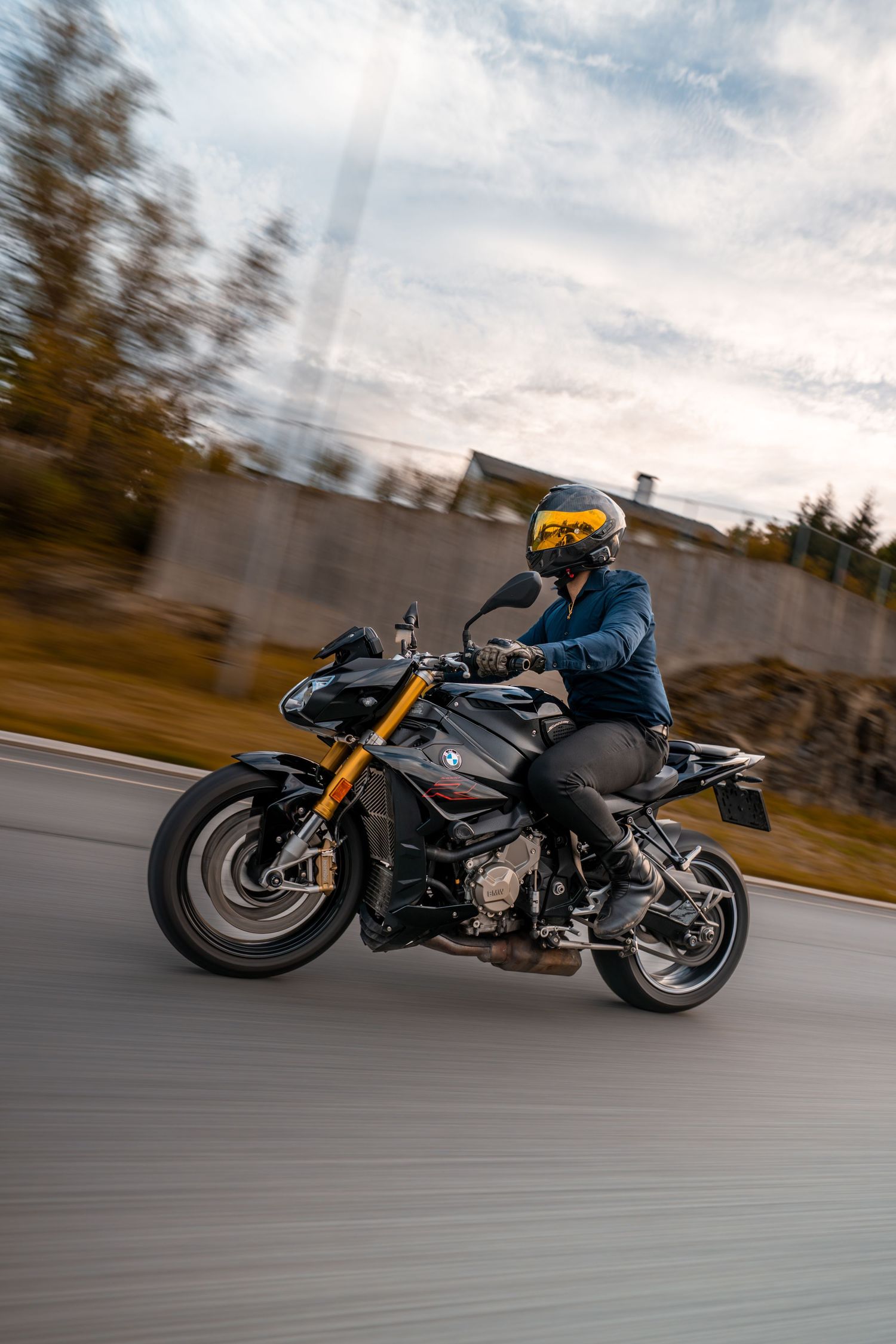 A motorcyclist in protective gear rides a sleek black sports bike with gold accents on a curving road at sunset.