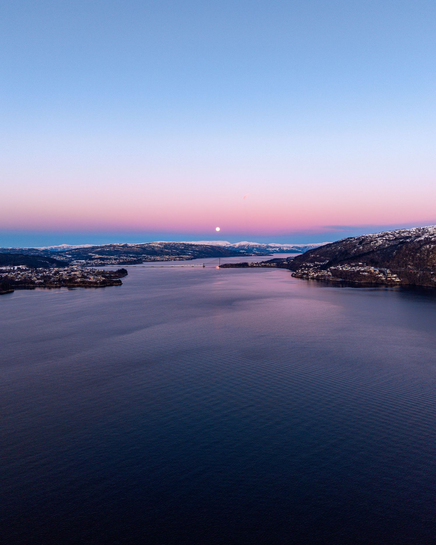 Aerial twilight view of a calm fjord with snow-dusted hills, pink horizon, and a full moon rising at dusk.