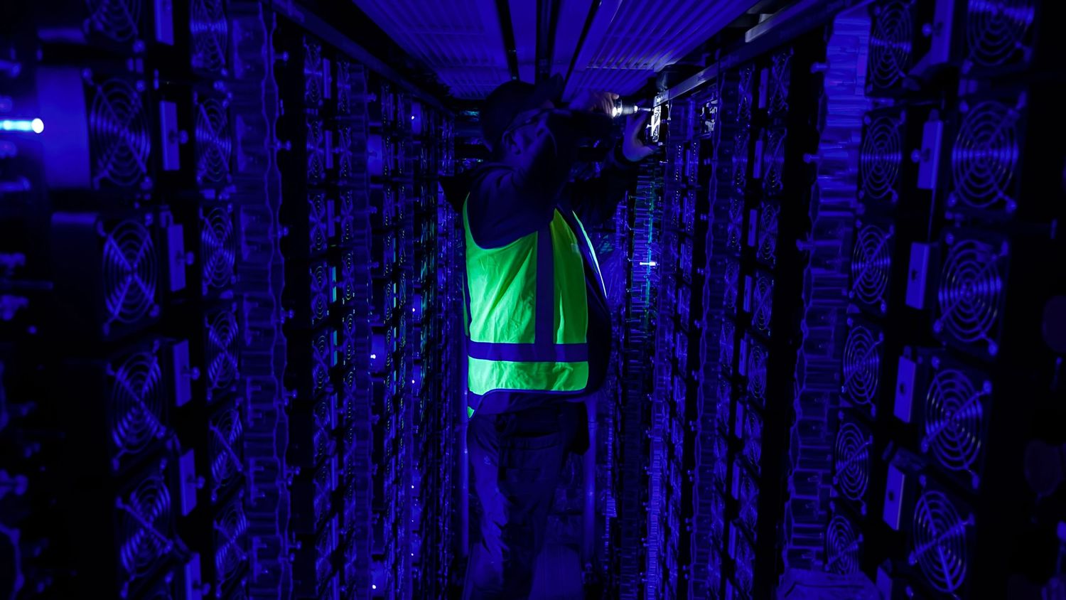 Technician in green safety vest working in a dark blue-lit server room data center.