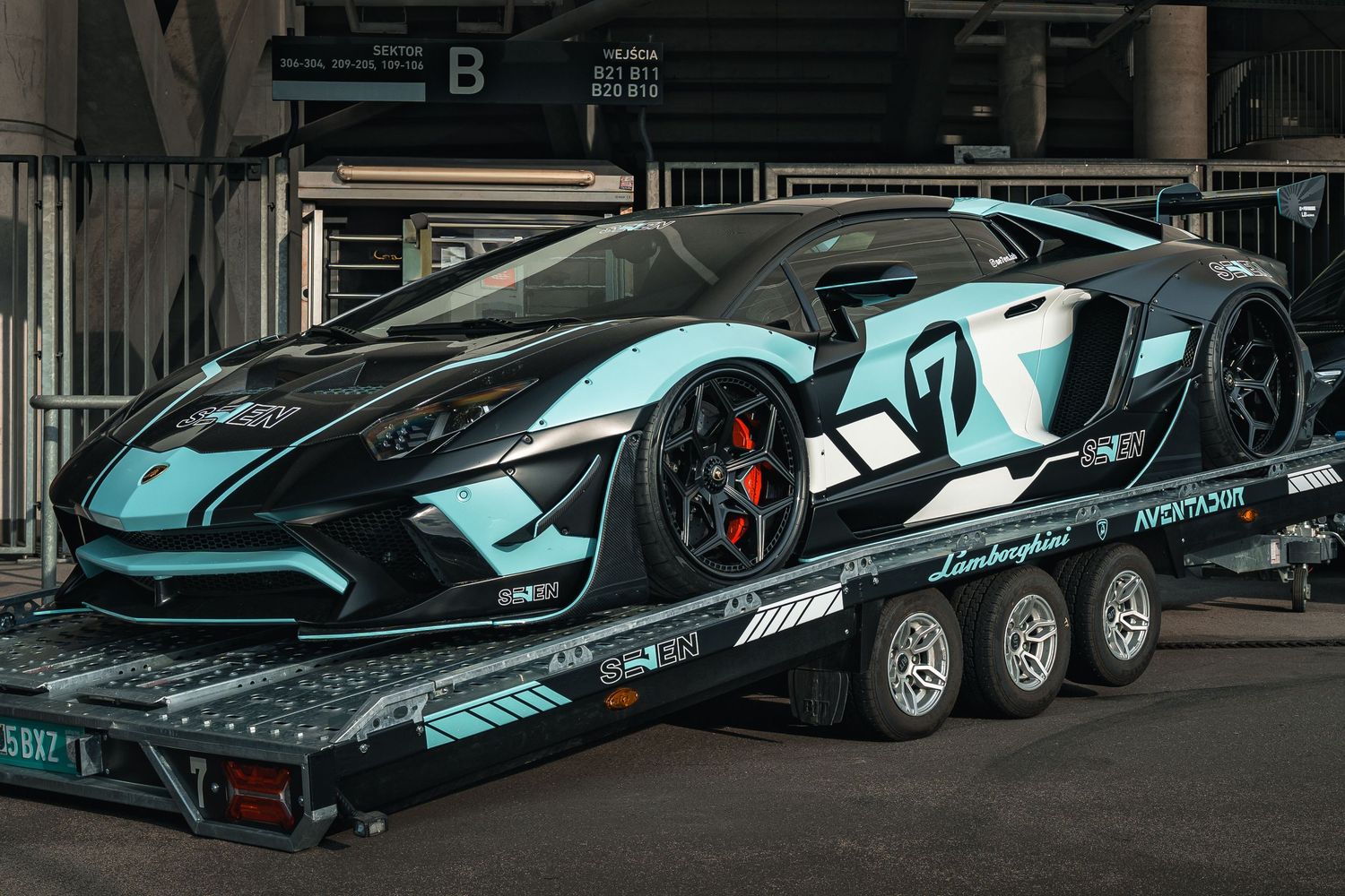 High-performance McLaren hybrid supercar in turquoise color scheme loaded on a black trailer in a dark garage setting.