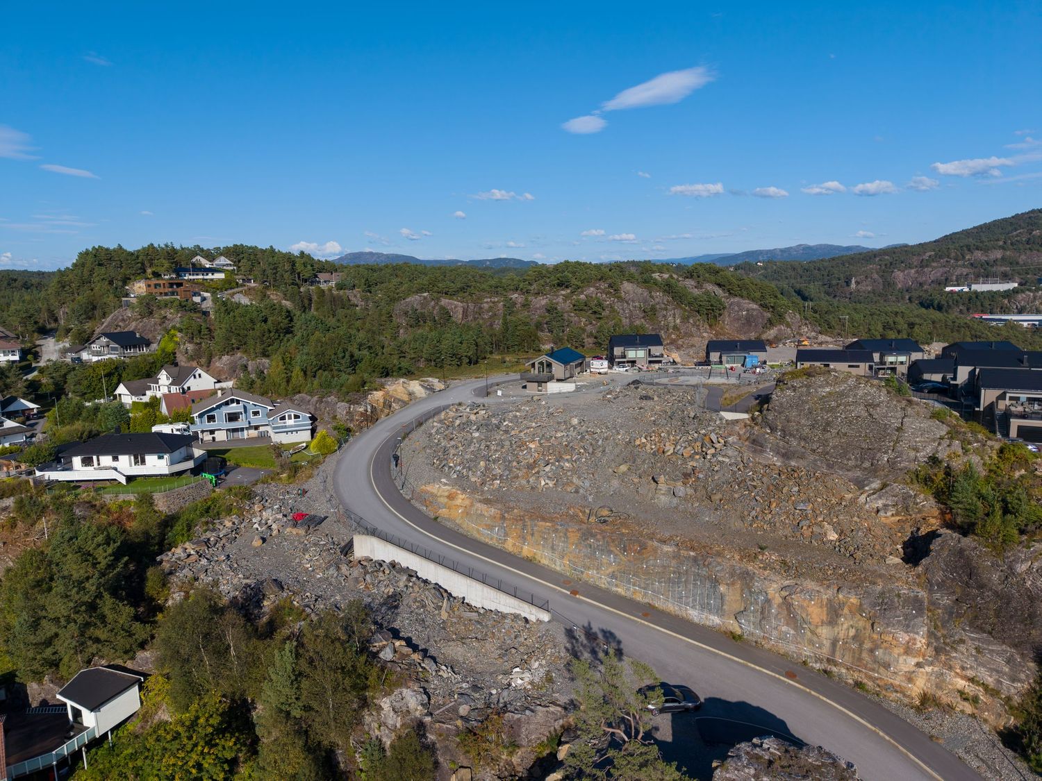 Aerial view of a curved road cutting through exposed rock face beside hillside homes with forested slopes under a blue sky.