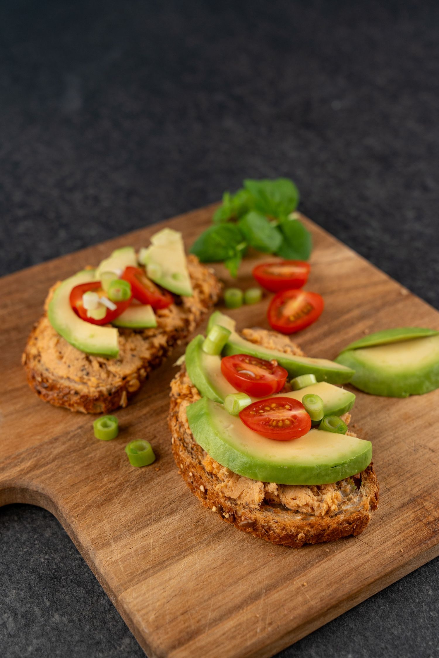 Two open-faced sandwiches on whole grain bread with spread, avocado slices, cherry tomatoes and green onions on a wooden board.