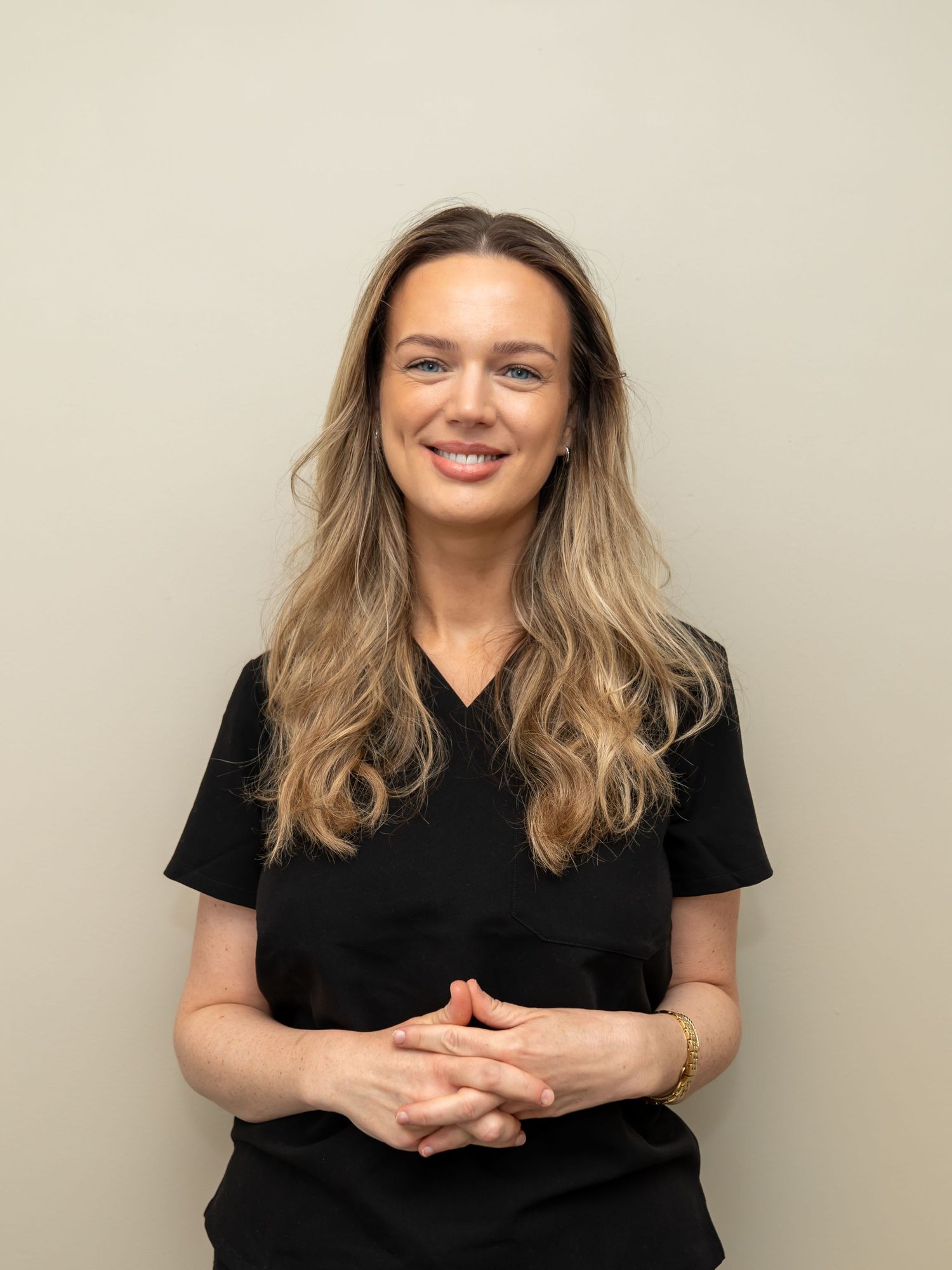 Smiling woman with long wavy blonde hair wearing black scrubs and a gold bracelet, standing with clasped hands against a neutral beige wall.
